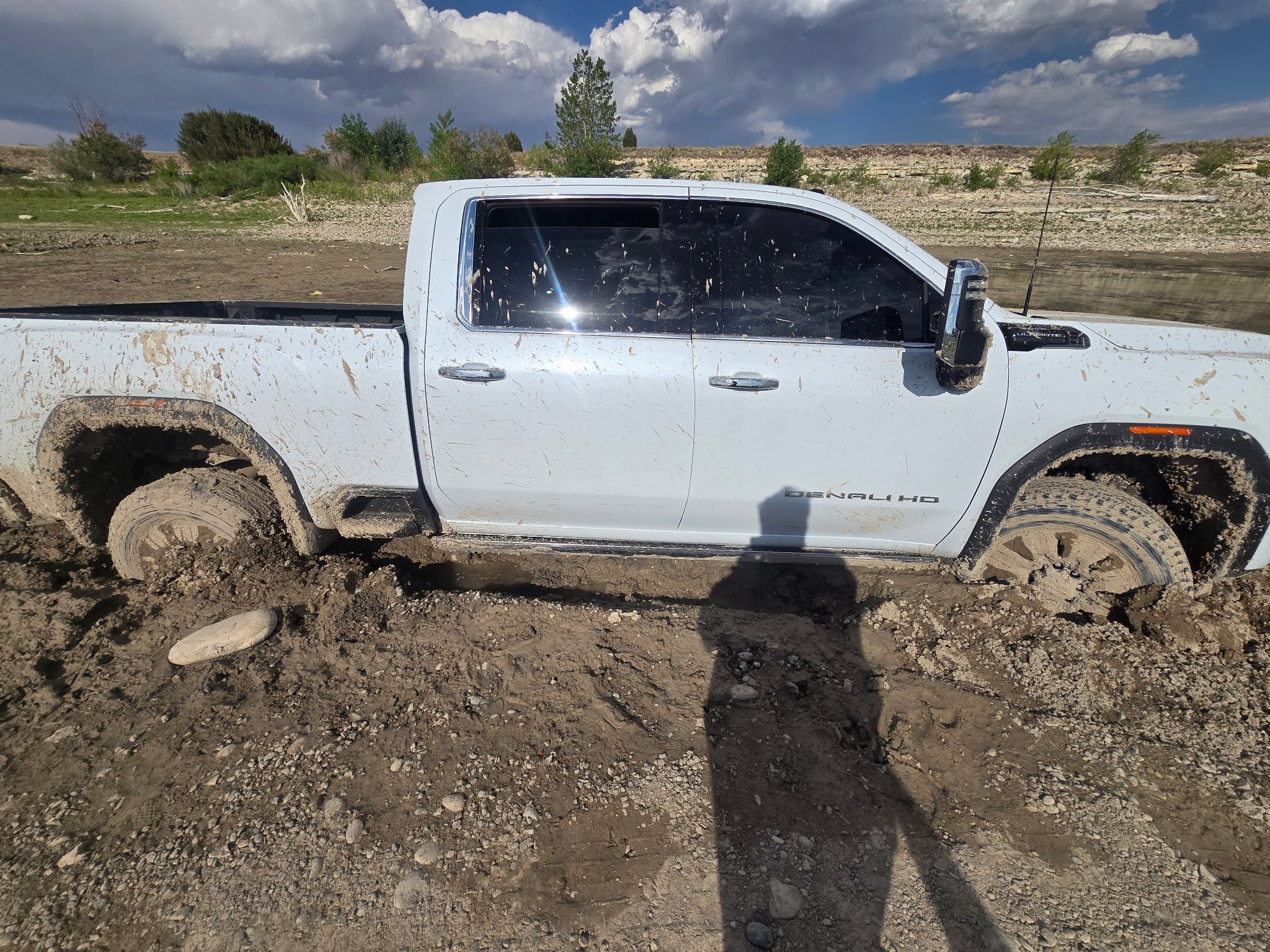 A white pickup truck with muddy tires and dirt splatters is parked on a rough dirt surface near a body of water, with a backdrop of trees and cloudy sky.