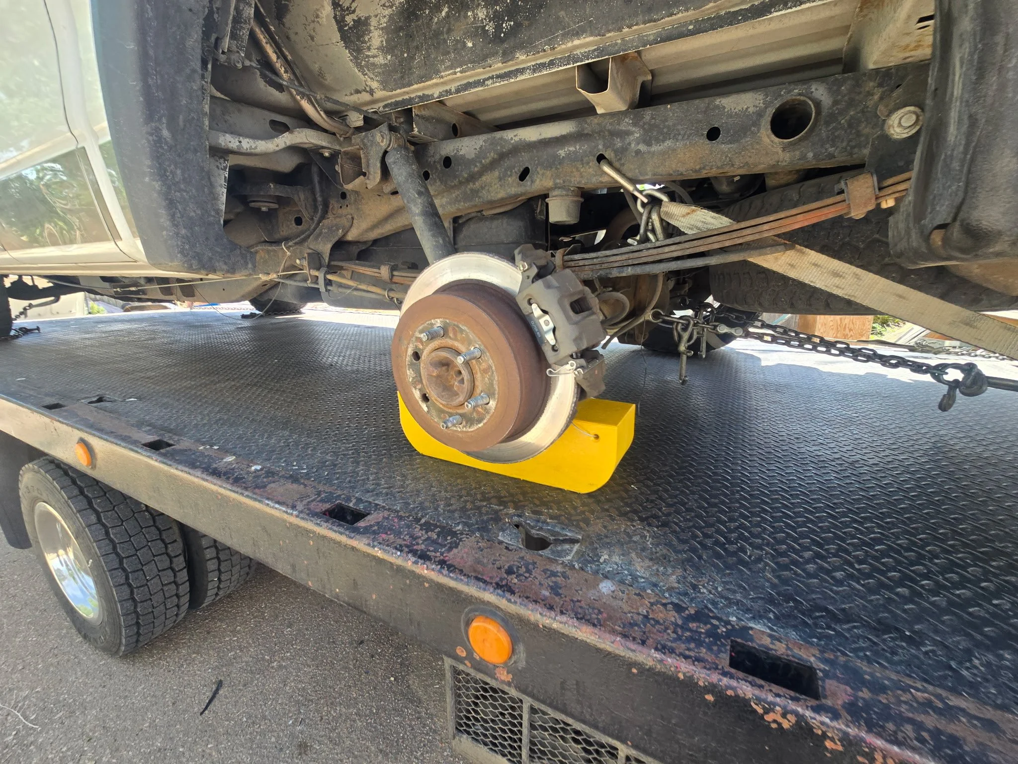 View of a flatbed tow truck with a rusty car wheel hub assembly and brake caliper, multiple chains securing the vehicle, and parts of the truck's bed, with some rust and dirt.