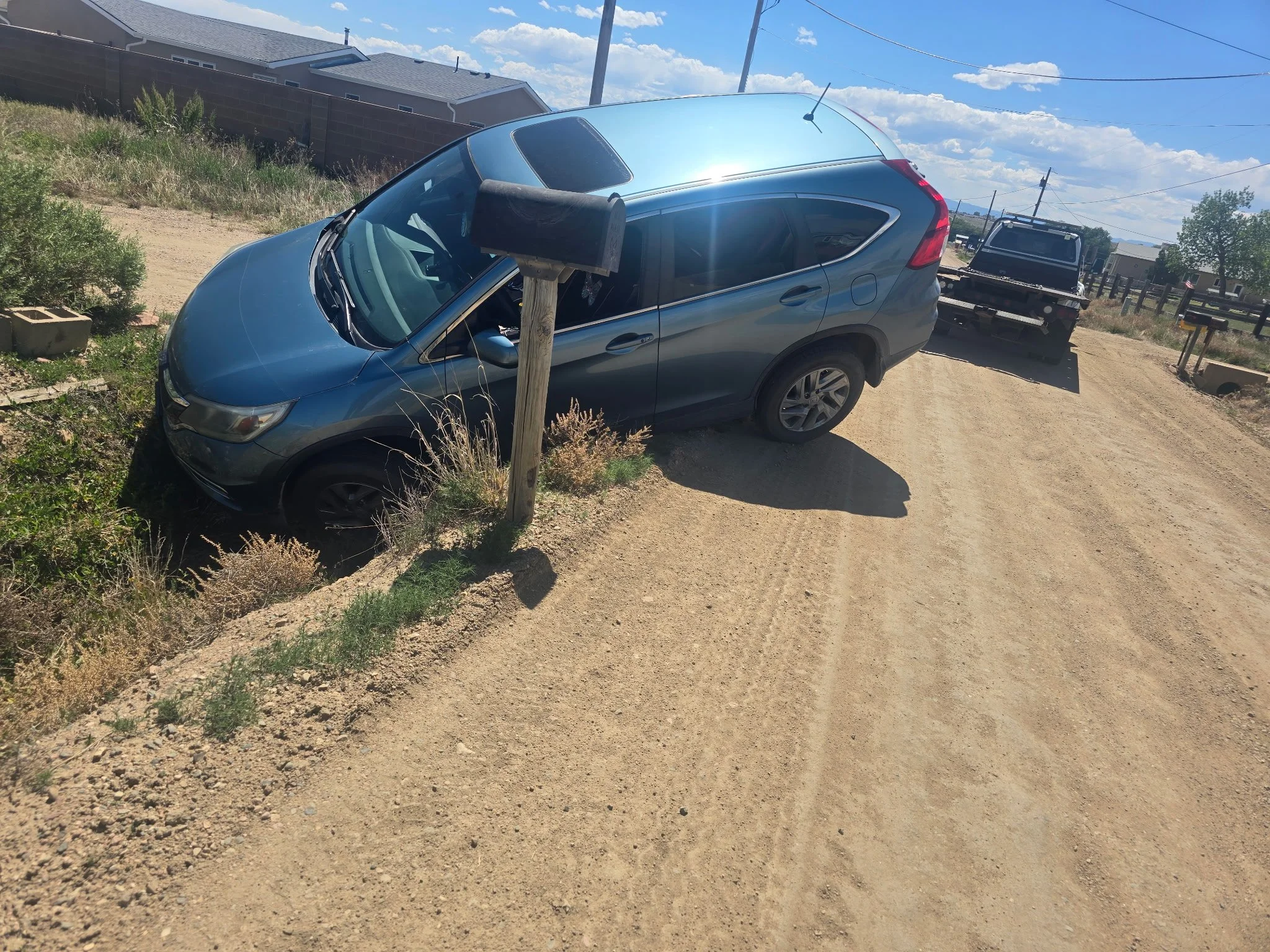 A blue SUV parked on a dirt road beside a wooden post with a mailbox, in a rural area with houses, fences, power lines, and clear skies in the background.