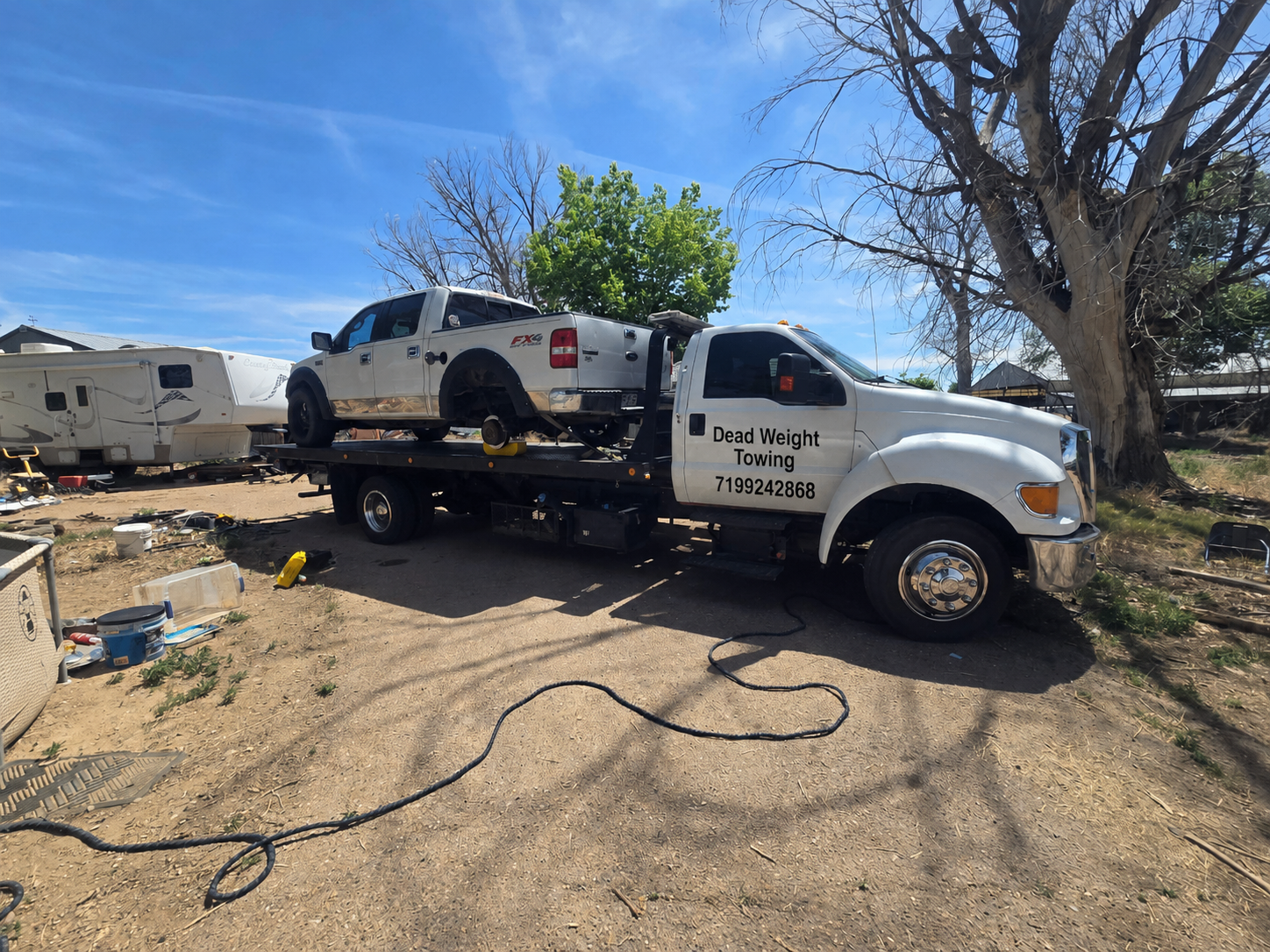 A flatbed tow truck labeled 'Dead Weight Towing' carrying a silver pickup truck with no wheels. The truck is parked on a dirt lot with various scattered items, including tools and a trailer. There are trees and a building in the background under a bl