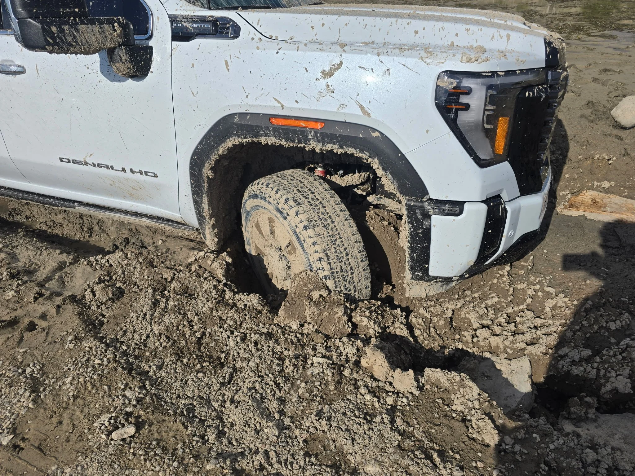 A white pickup truck with mud splatters on the side and tire, parked on a dirt ground with mud and rocks. The front part of the vehicle includes a black grille, headlights, and a large side mirror.
