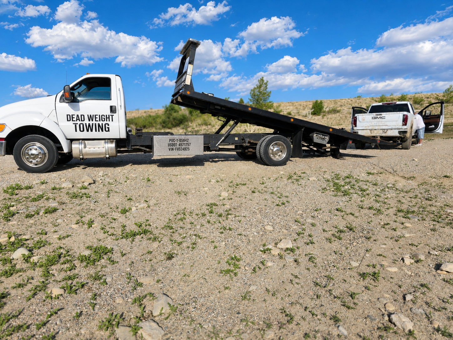 A flatbed tow truck labeled 'Dead Weight Towing' is towing a white pickup truck on a gravel lot under a blue sky with scattered clouds. A person is working near the passenger side of the pickup truck.