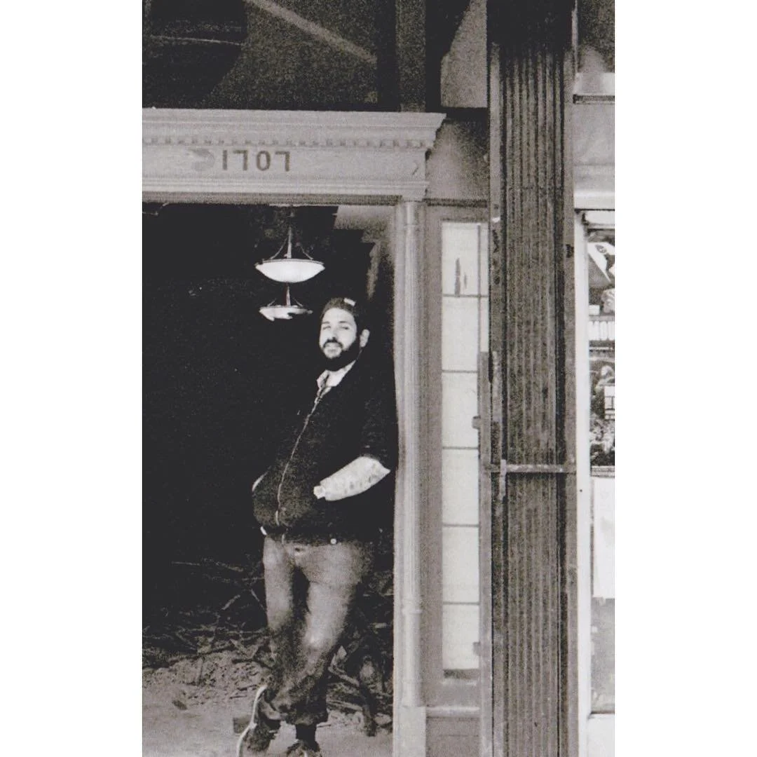 A man with a beard and tattoos leaning against a wall outside of a building with the number 1107. The photo is in black and white.