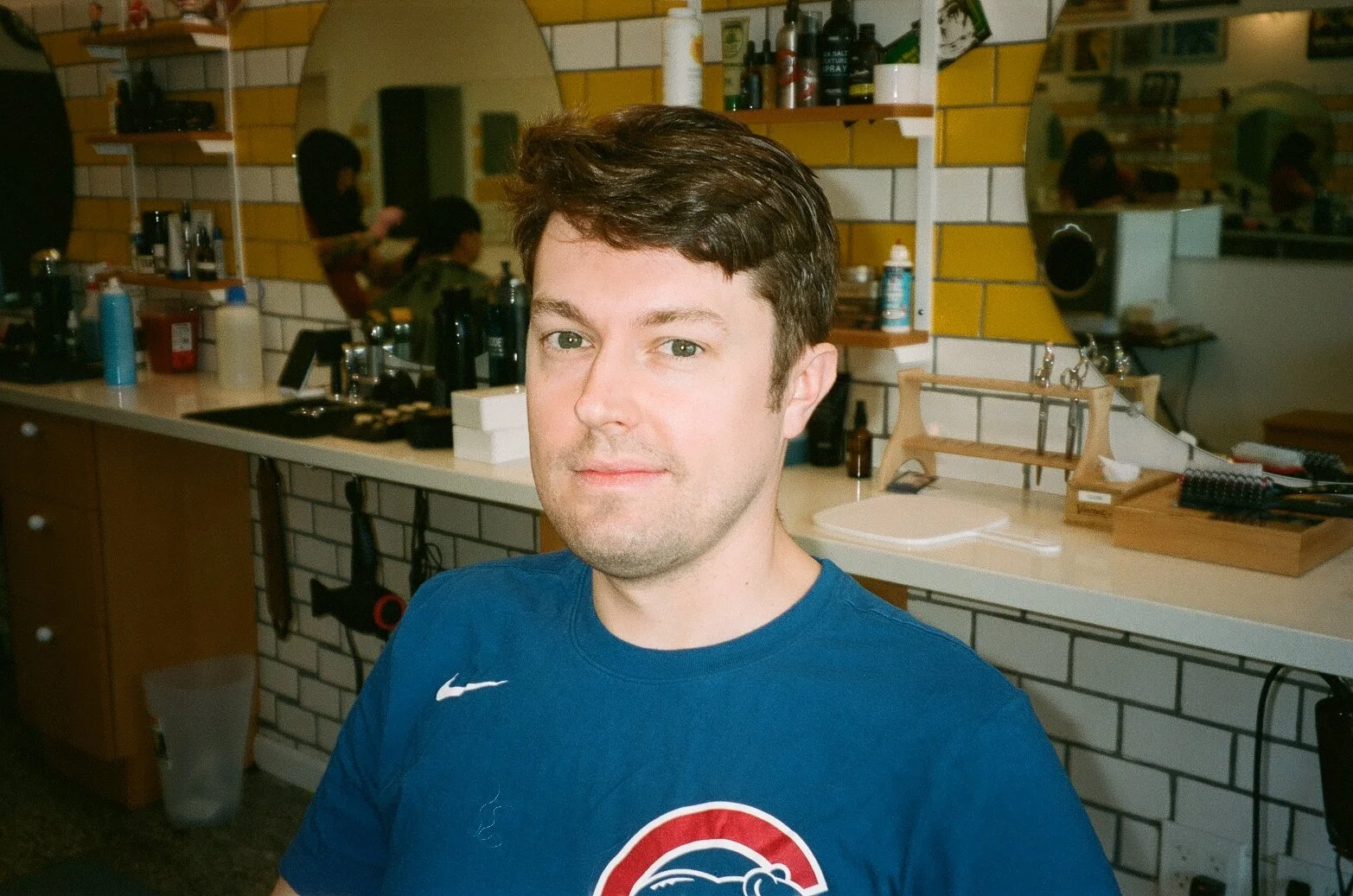 A young man with short brown hair and light skin wearing a blue Nike t-shirt with a Chicago Cubs logo, sitting in a barber shop or hair salon. The background shows shelves with styling products and tools, a mirror, and other people receiving haircuts