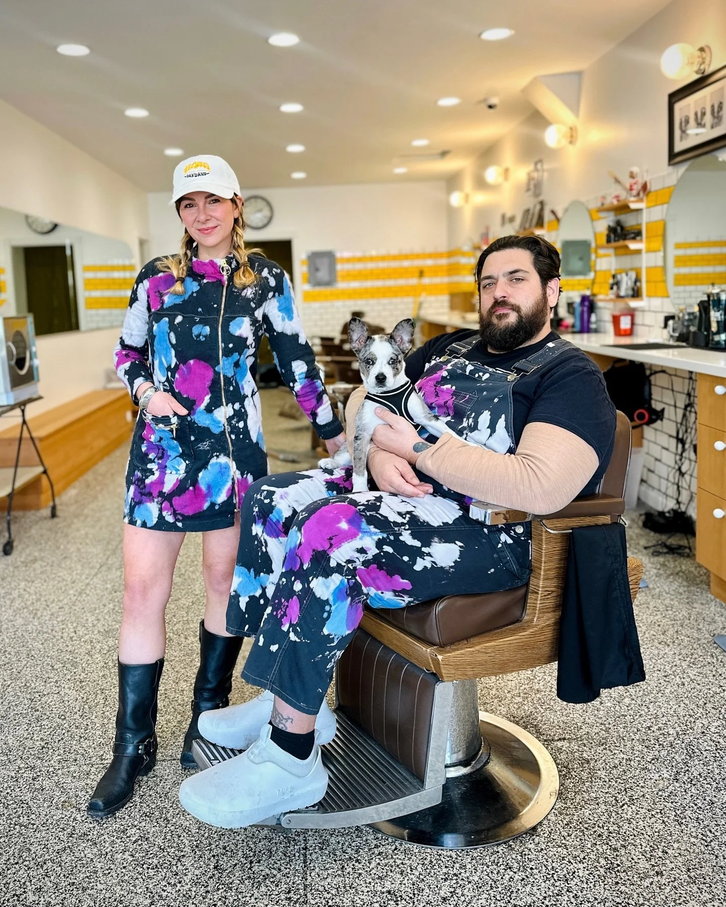A woman is standing and smiling next to a man sitting in a barber chair, holding a dog. They are in a retro-style barbershop with bright lighting and yellow accents on the walls.
