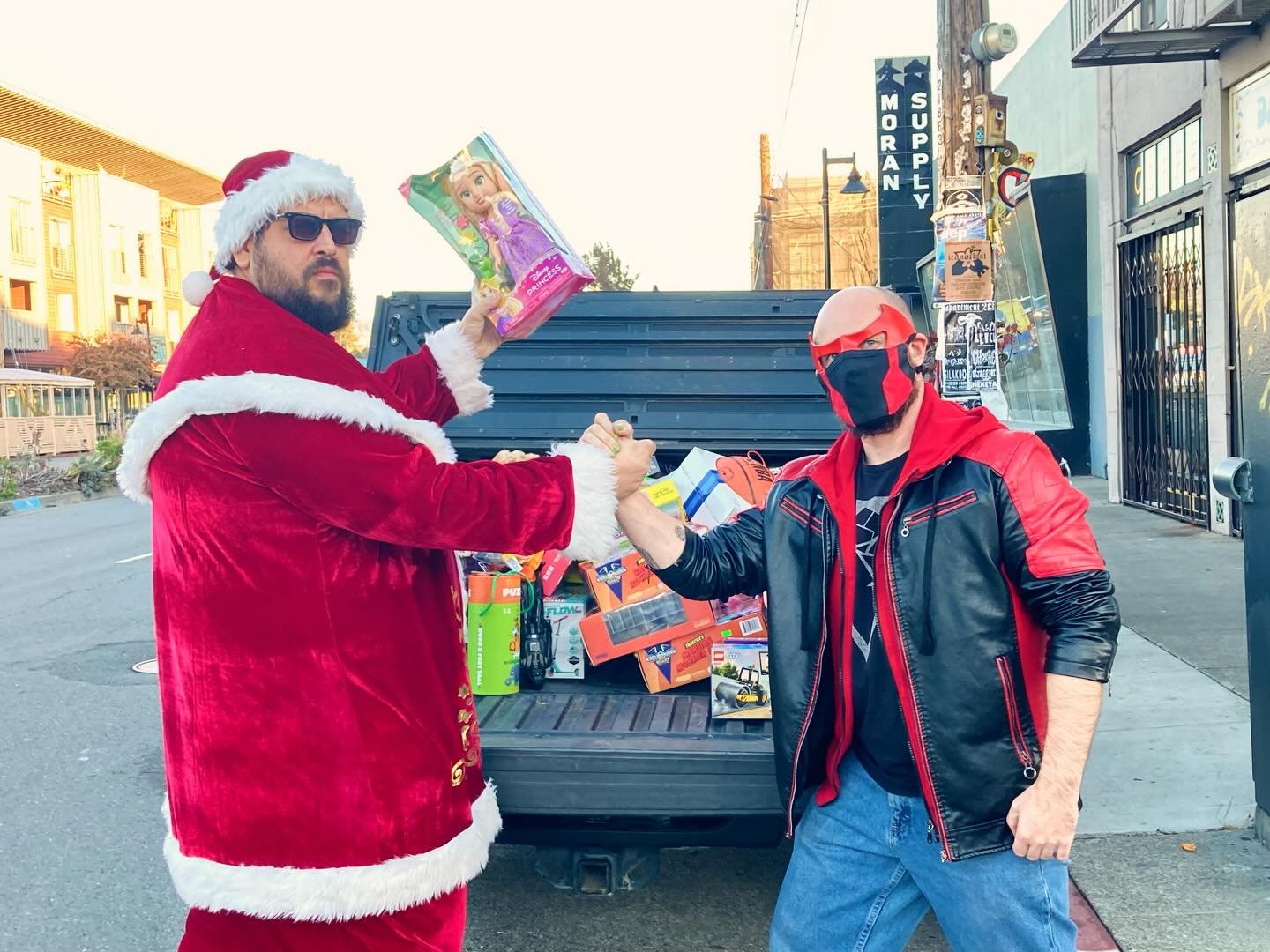 Two men in front of a truck filled with toys, shaking hands. One man is dressed as Santa Claus with sunglasses, a Santa hat, and a red velvet suit. The other man is wearing a black and red leather jacket, jeans, a face mask, and sunglasses.
