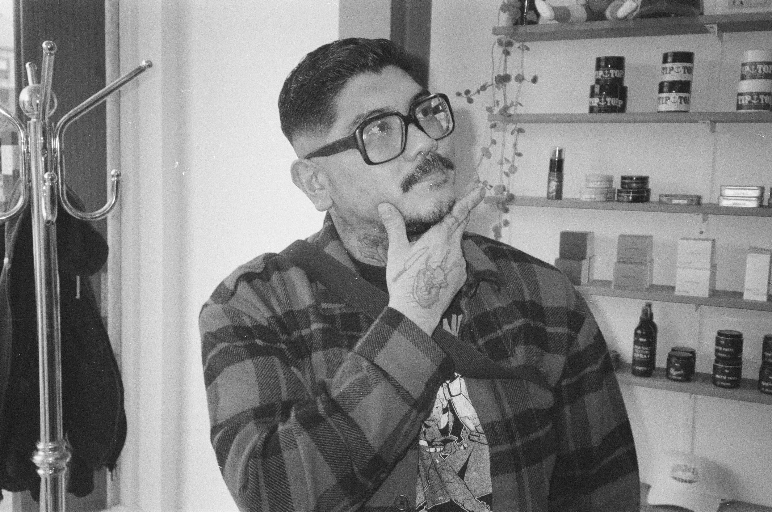 A man with slicked-back hair and a mustache wearing glasses and a plaid shirt, touching his chin with his hand, sitting or standing in a room with shelves and various grooming products.
