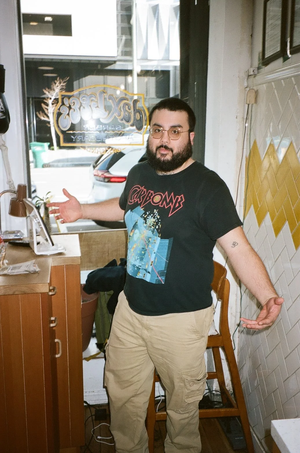 A man with a beard and glasses standing inside a coffee shop, gesturing with his arms open. He's wearing a black graphic T-shirt and khaki pants. Behind him is a window showing parked cars outside.