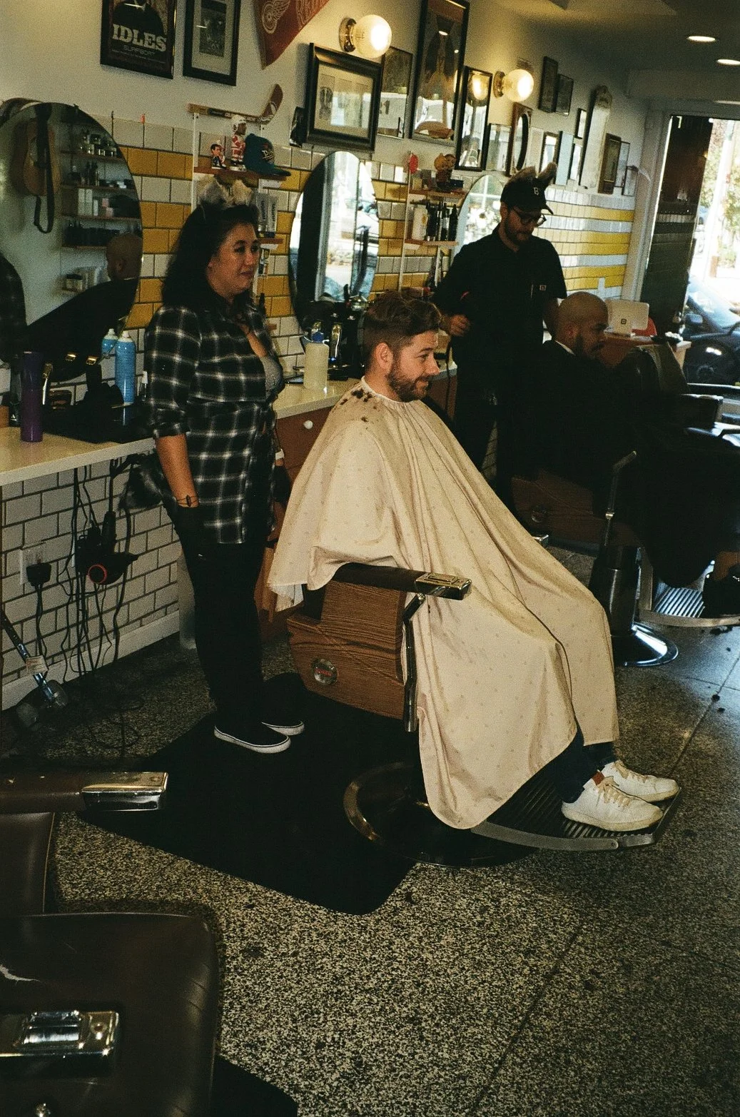 A man receives a haircut at a barbershop while seated in a vintage barber chair, with a barber and another customer nearby, and a stylist or barber standing behind him. The shop has framed pictures on the wall, mirrors, and various barber tools on th