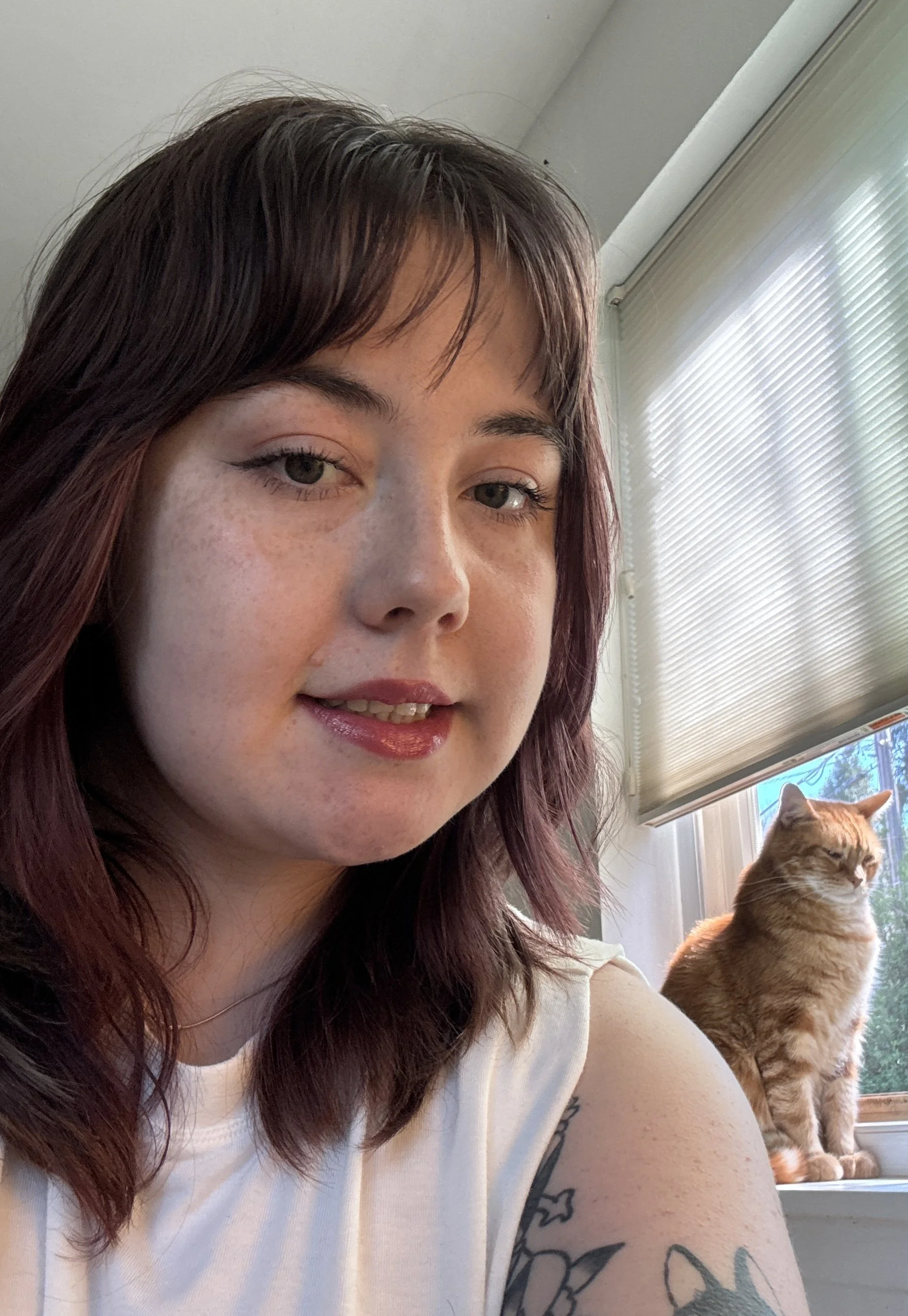 A close-up of a woman with dark red hair and tattoos on her arm, smiling slightly, sitting by a window with blinds, with an orange tabby cat sitting on the window sill.