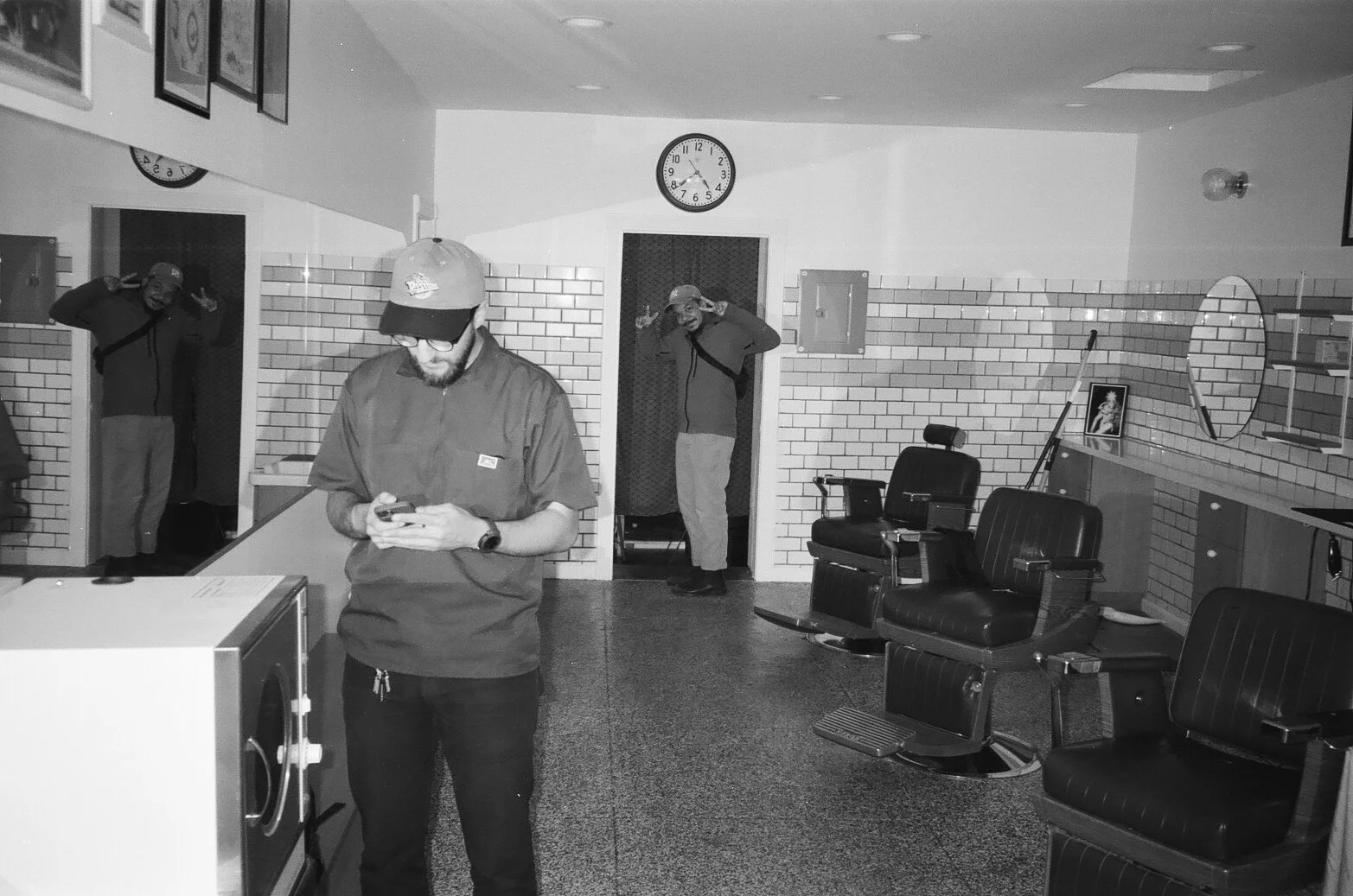 An indoor barbershop with retro decor, featuring black barber chairs, a wall mirror, and framed picture on the wall. Three men are in the shop: one in the foreground looking at his phone, and two in the background near doorways, making peace signs wi