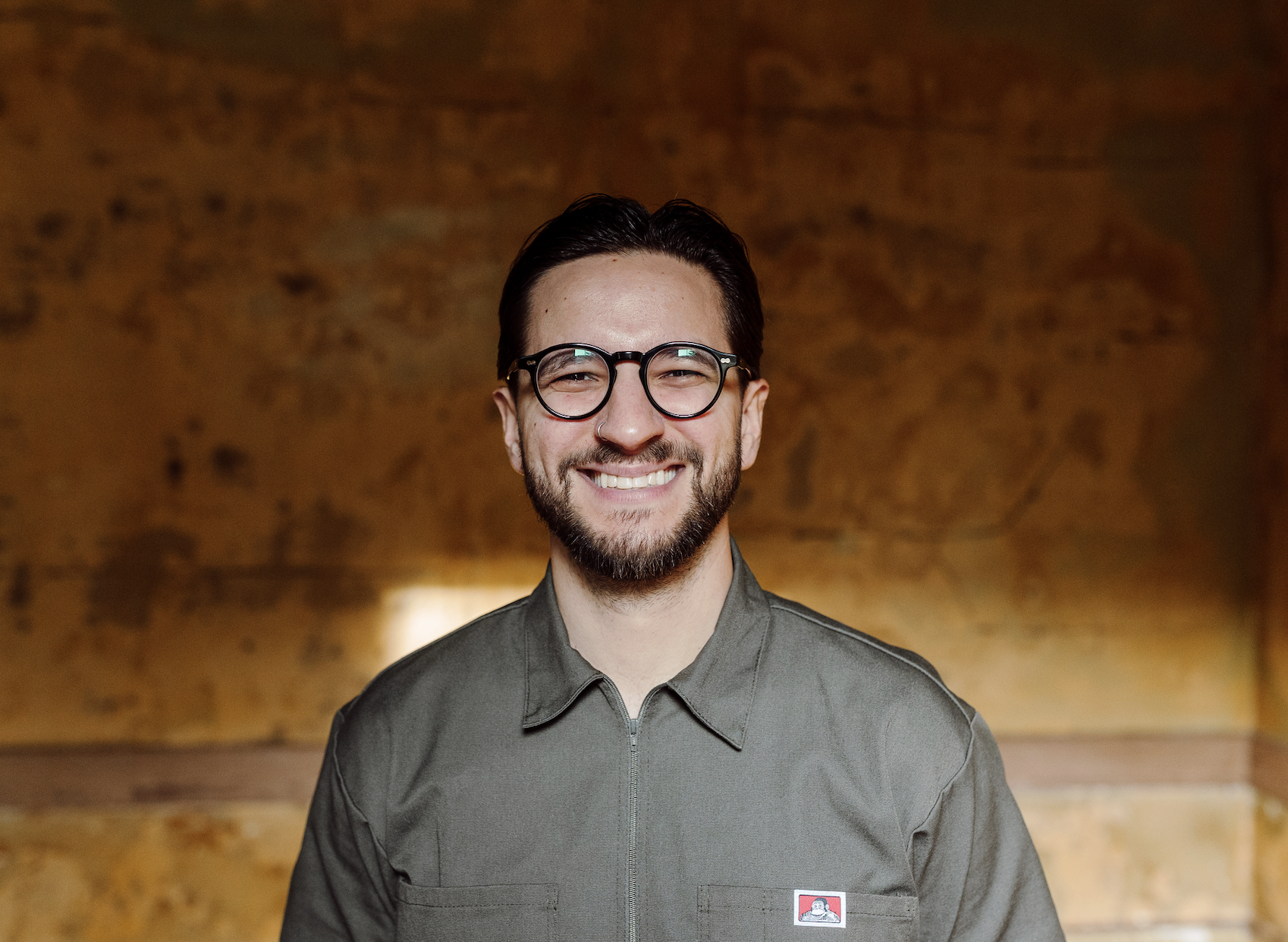 A smiling man with glasses and a beard, wearing a gray work shirt with a logo, standing against a stone wall background.