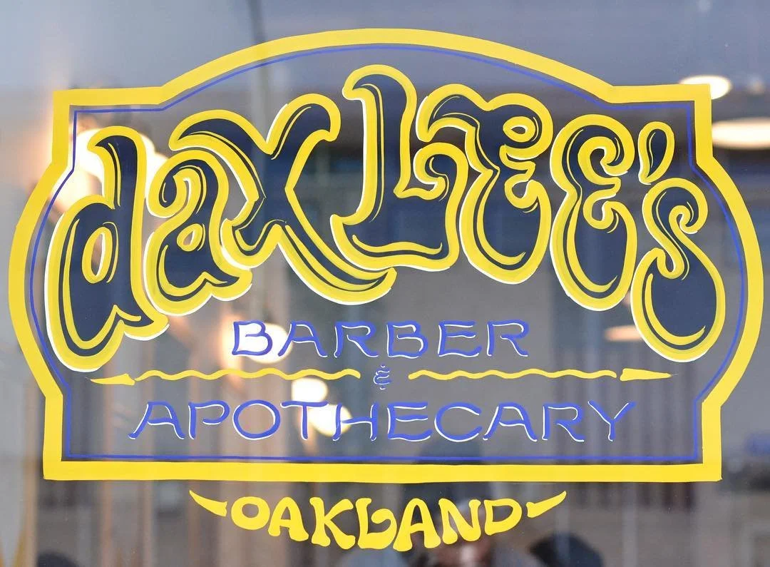 Sign for Clint Liles Barber & Apothecary in Oakland, with yellow and blue lettering on a transparent window.