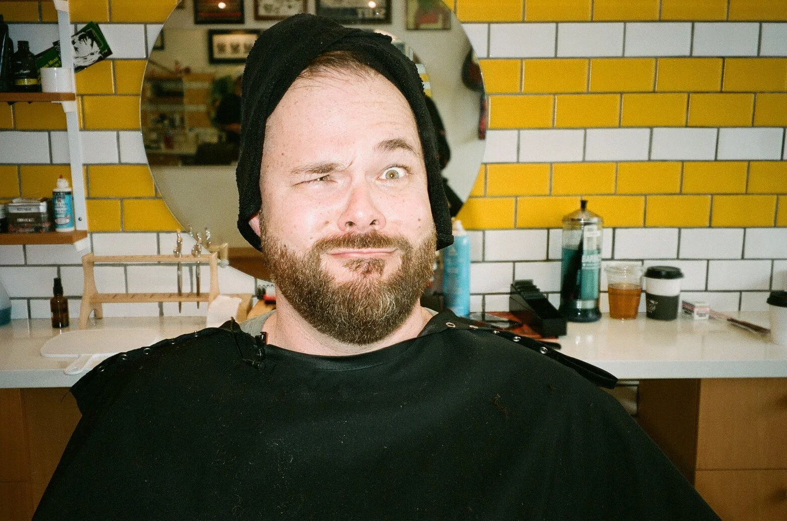 A man with a beard and mustache, winking and smiling, wearing a black hat and clothing, sitting in a barbershop or salon with a colorful brick wall and various supplies in the background.