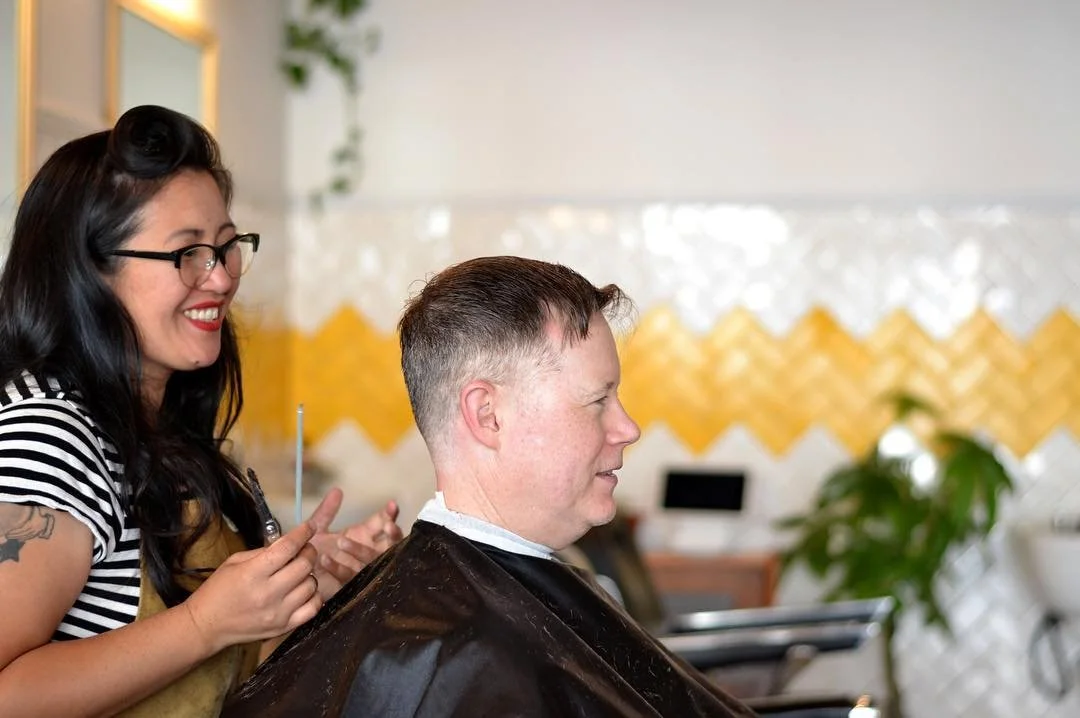 A woman with glasses and long black hair getting a haircut from a smiling hairstylist with glasses in a salon.