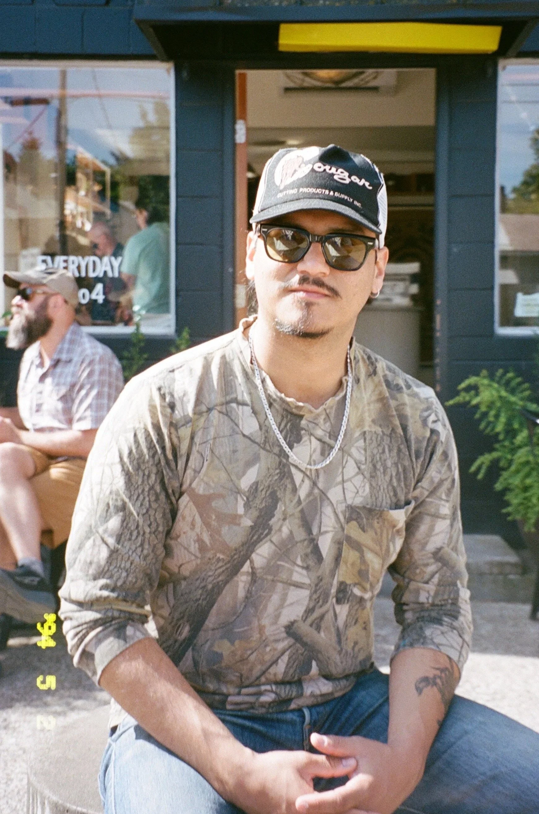 A man wearing sunglasses and a camouflage shirt posing outdoors.