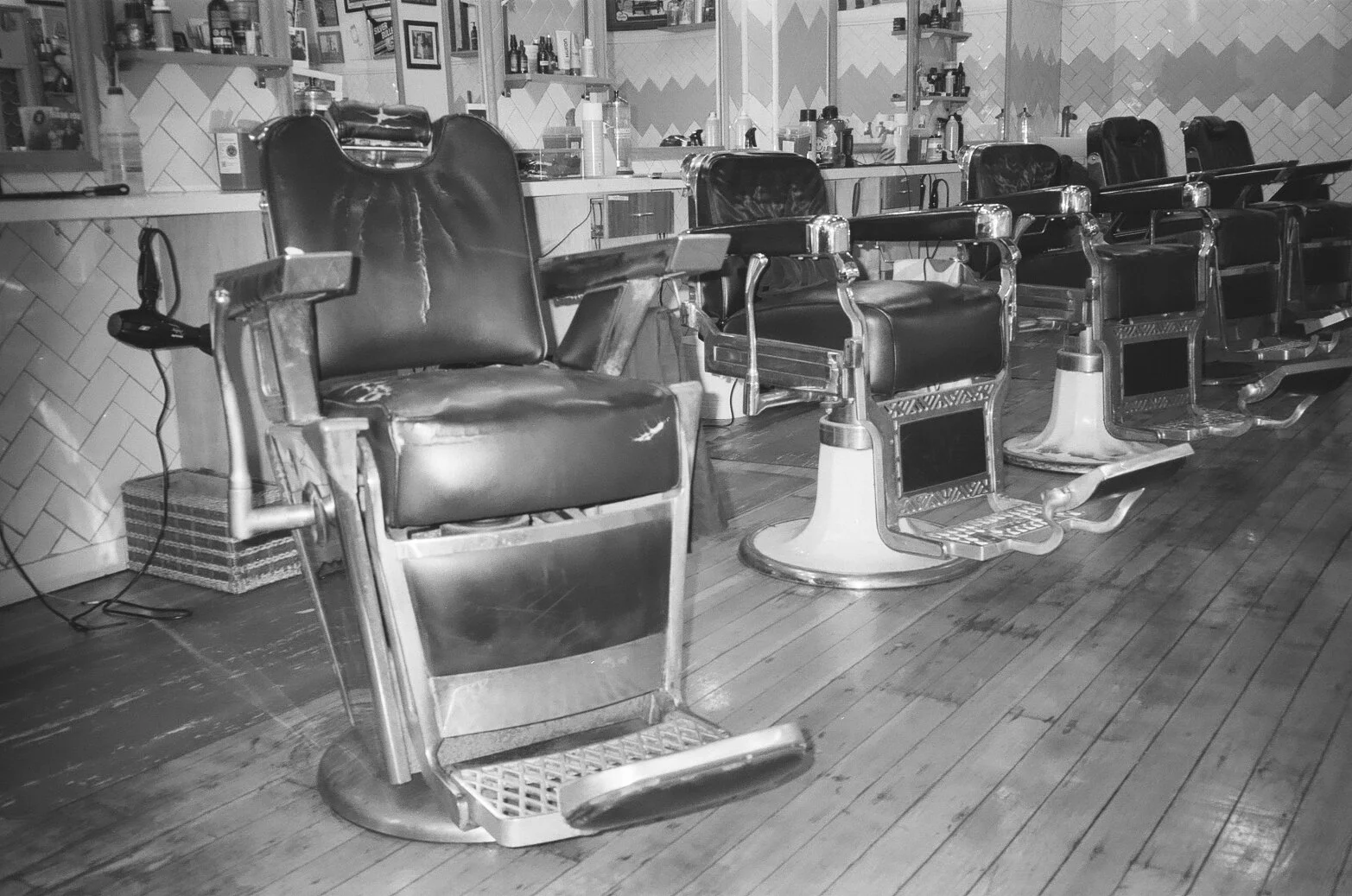 Old vintage barber shop chairs lined up in a row in a barbershop with a wooden floor and tiled wall in the background.