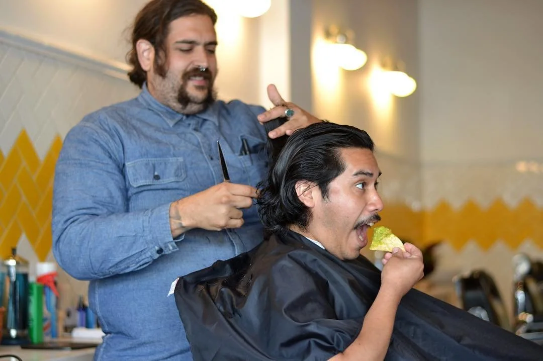 A man with black hair and a mustache eating a slice of lime-green cake or pie while seated in a barber's chair. A barber with dark hair and a beard, wearing a blue shirt, is standing behind him, holding scissors, and appears to be cutting or styling 