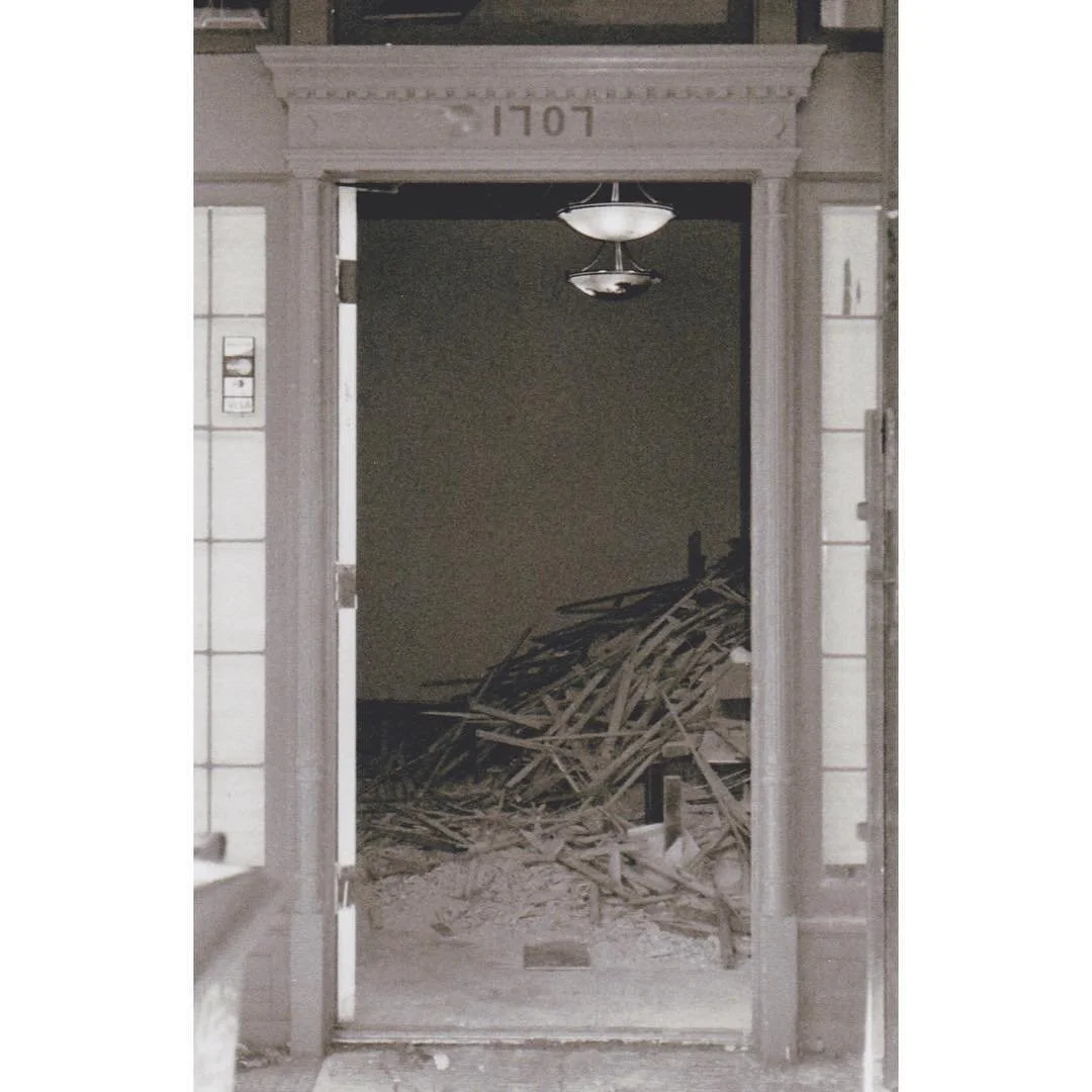 Dark doorway of a building with wood debris piled inside, illuminated by a hanging ceiling light, with a stone arch and the number 1707 above the entrance.