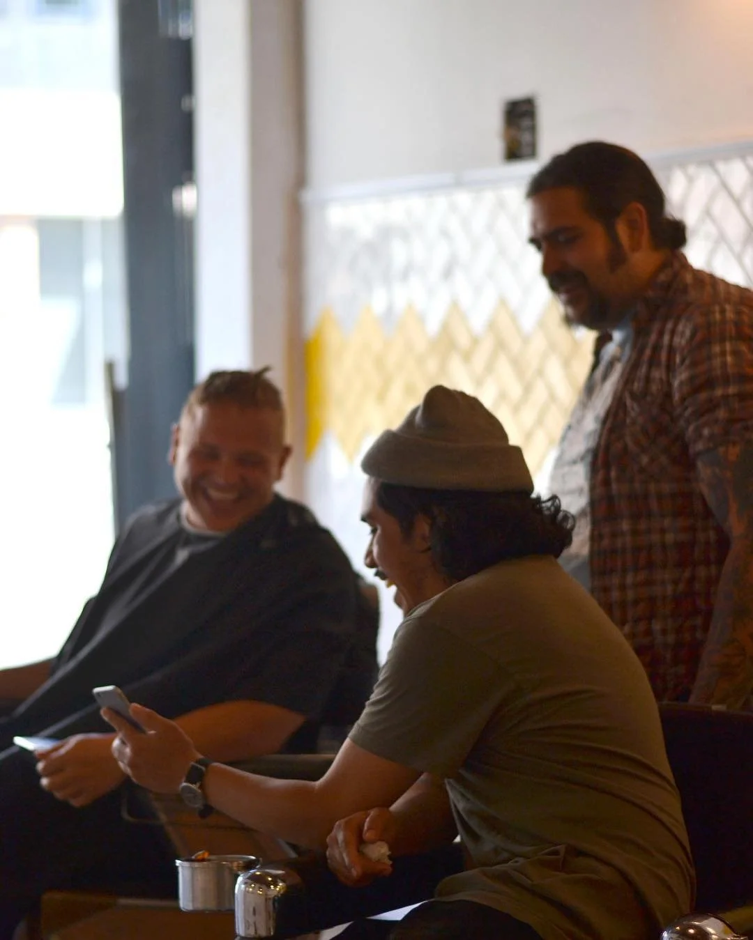 Three men are sitting and standing in a room, smiling and laughing, one using a phone