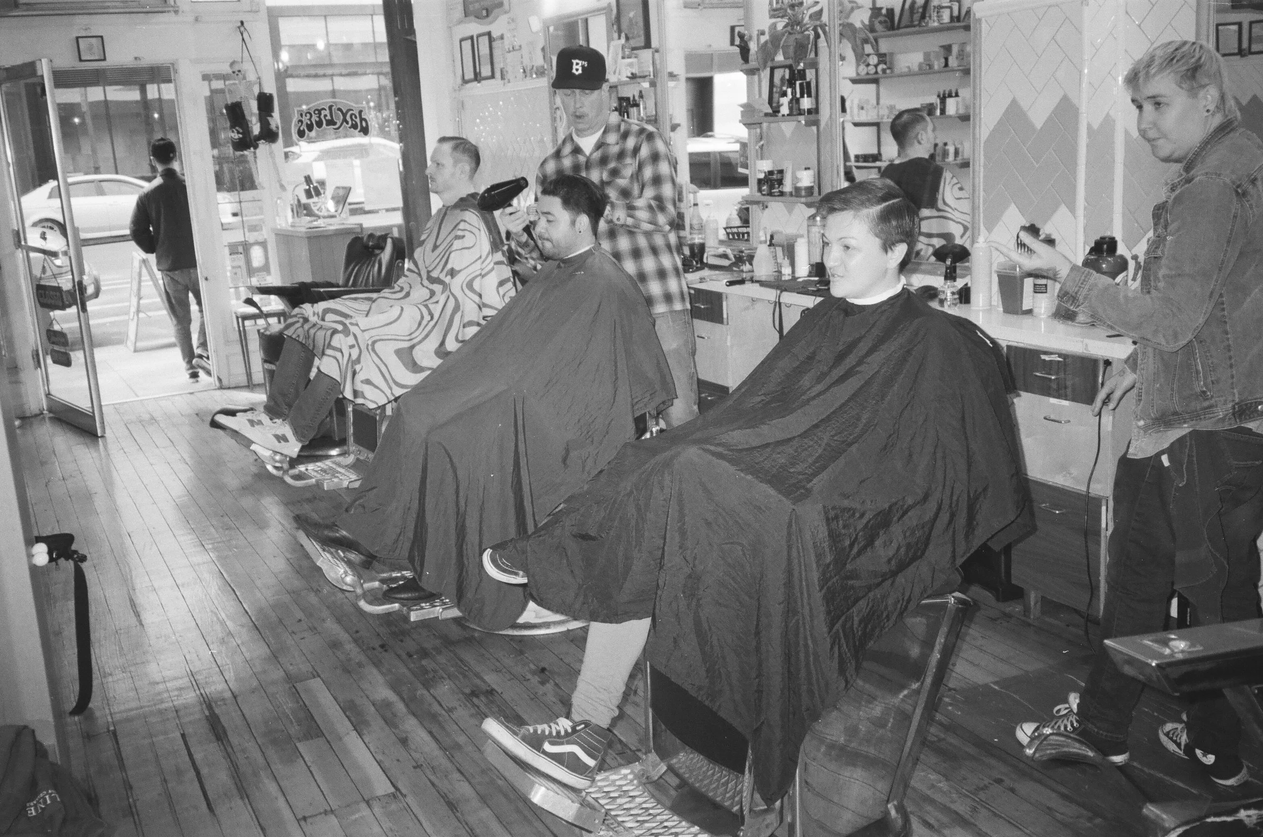 A black and white photo showing a barbershop with two people getting haircuts and a barber styling one customer's hair. One customer is seated in a barber chair with a barber working on his hair, and another customer is sitting in a barber chair with