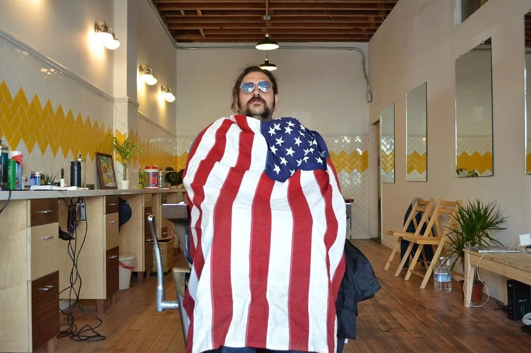 A man sitting in a chair in a salon, draped with an American flag and wearing sunglasses.