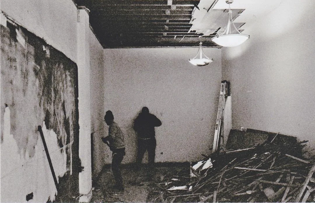 Two people inspecting a room with significant damage, including a collapsed ceiling with exposed wooden beams and debris scattered on the floor. The room appears to be under renovation or damaged from a structural failure.