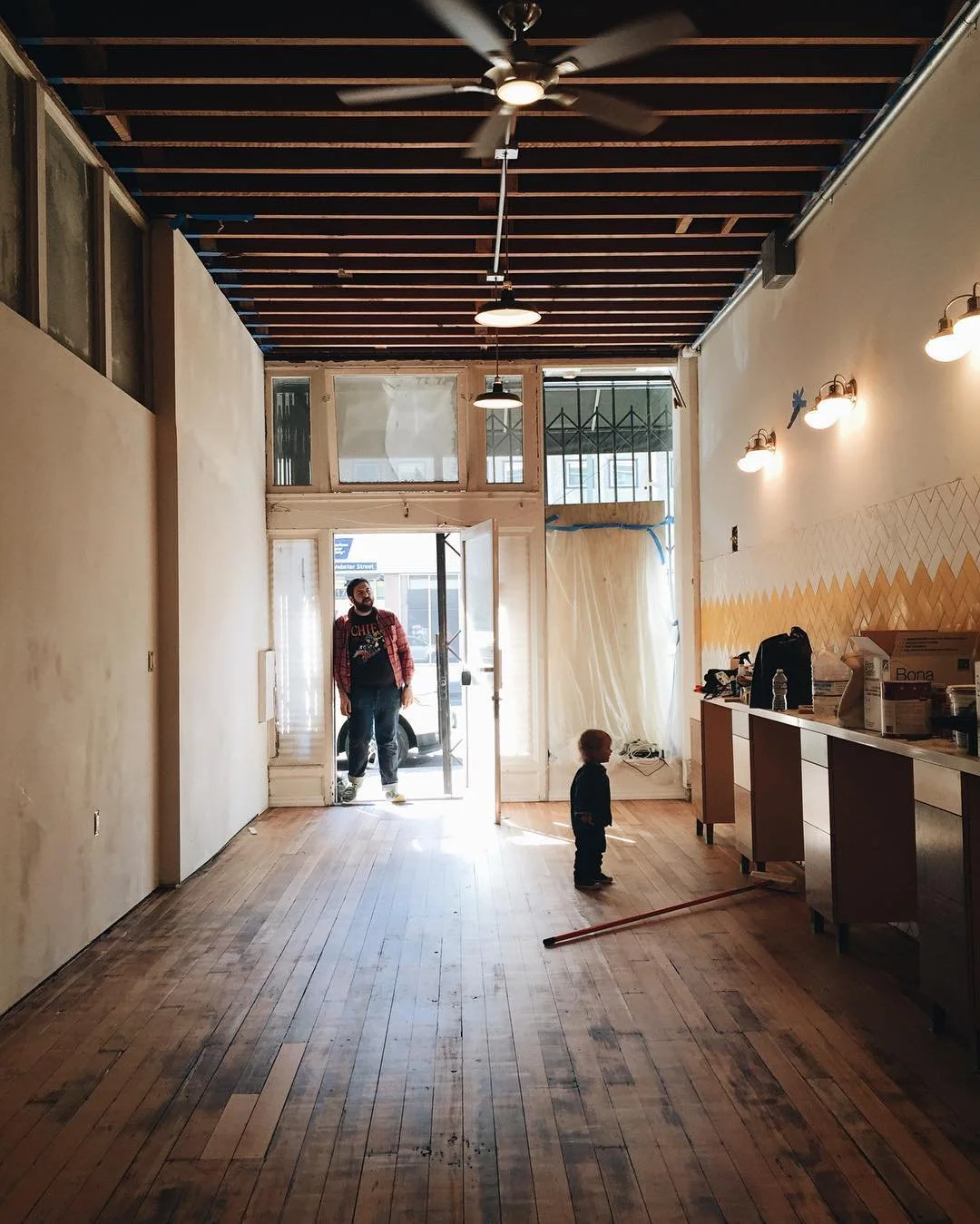 A spacious, partially renovated interior space with wooden floors, unfinished walls, exposed ceiling beams, and a large front window with a glass door. A man and a small child stand near the entrance, with the man holding the door open. There are wal