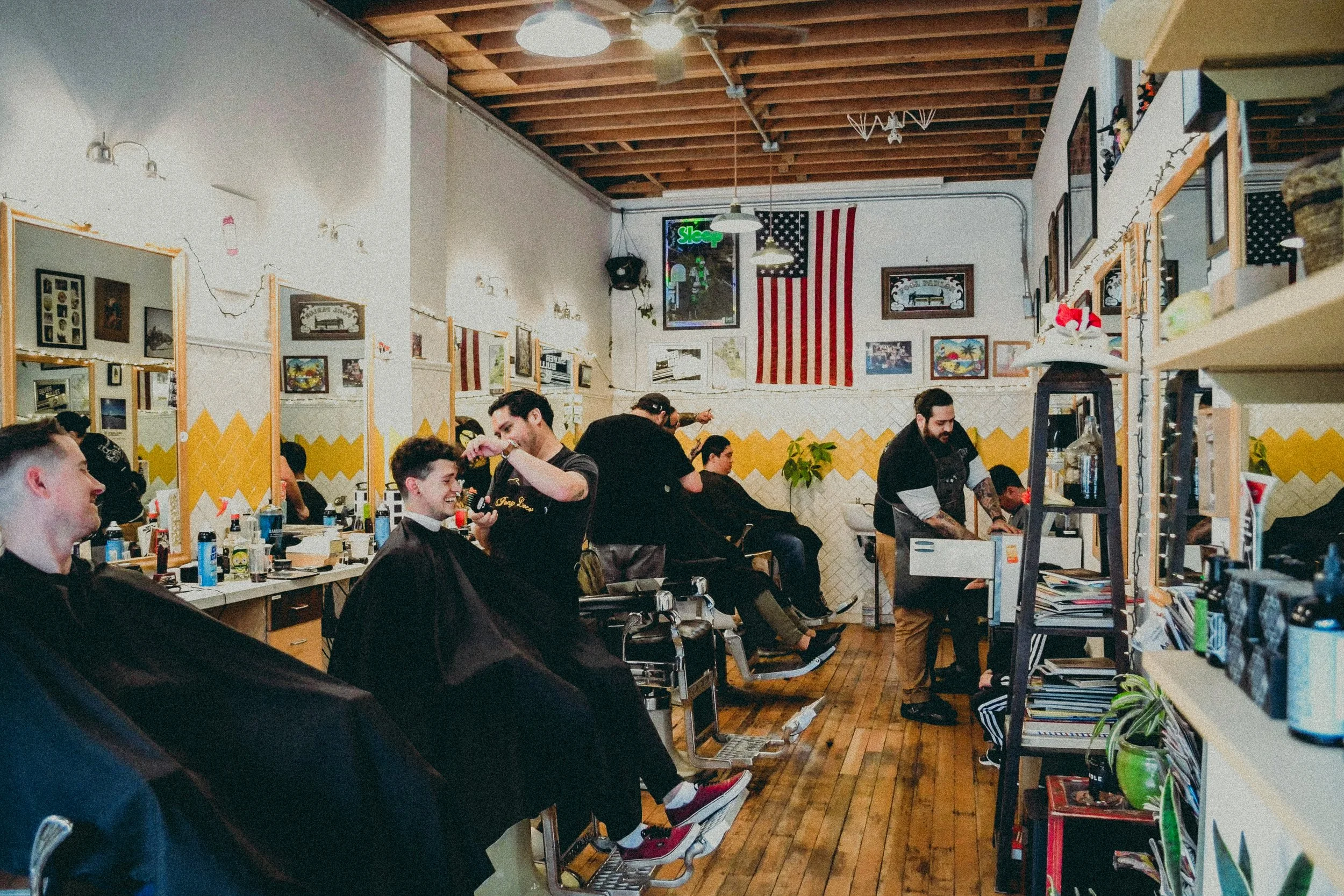 Barbershop with multiple clients getting haircuts and hair styling, barbers and customers engaging, and decorations including flags on the wall.