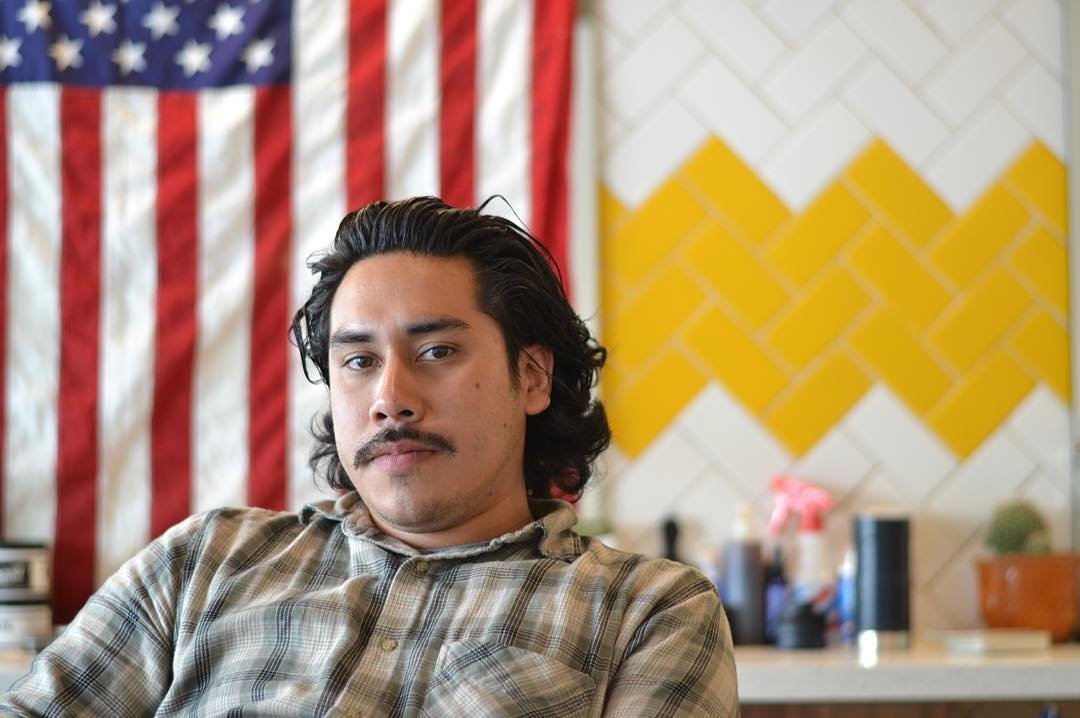 A young man with dark wavy hair and a mustache sits in a room with a large American flag and yellow and white painted wall behind him.