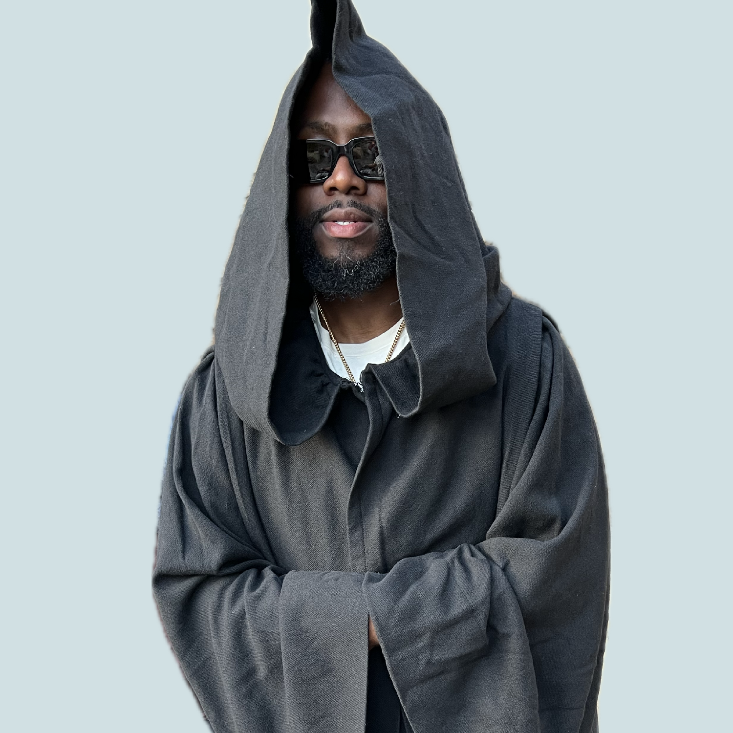 A man wearing black sunglasses and a black hoodie with the hood over his head, standing against a light gray background.