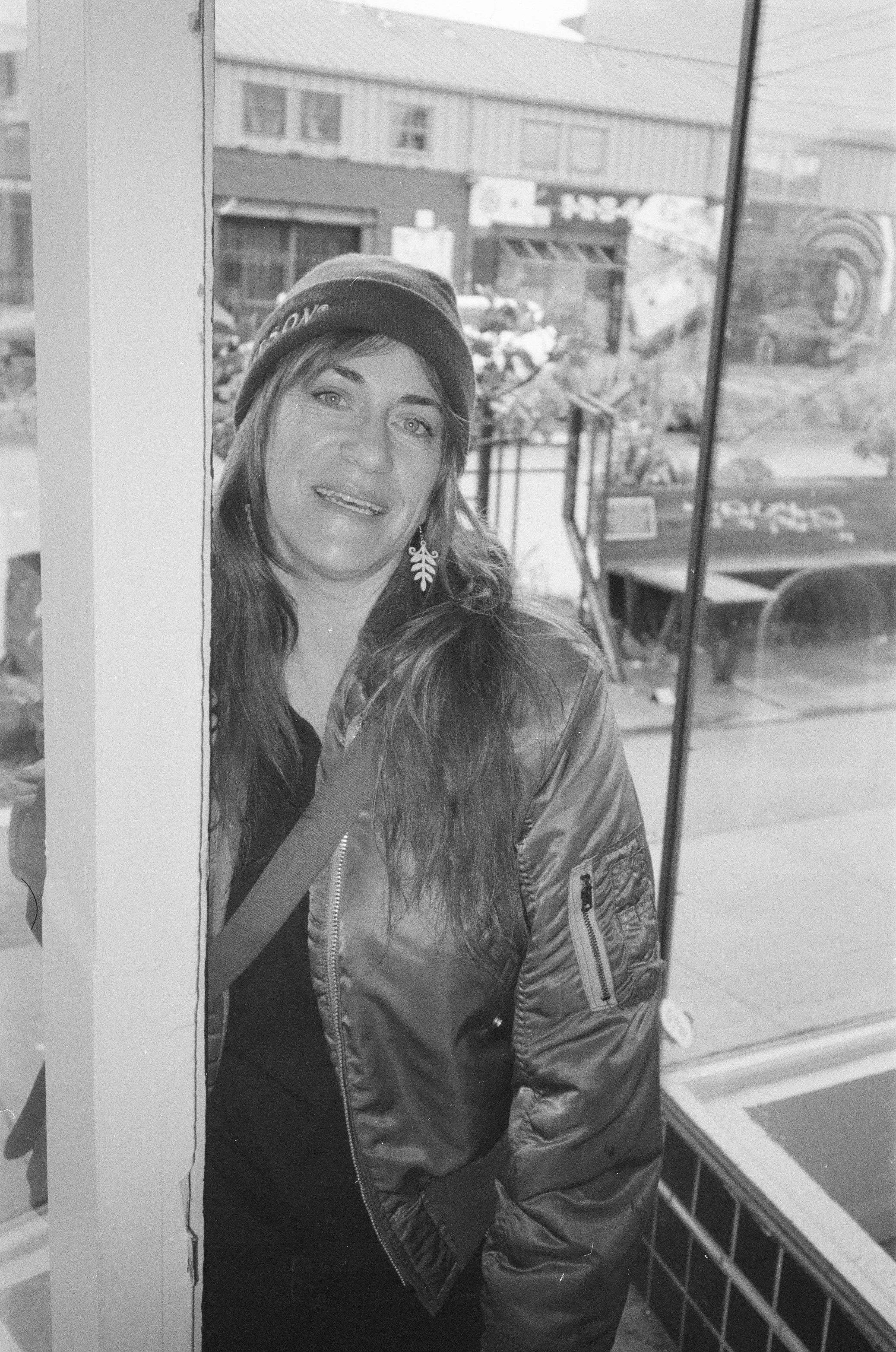 A woman with long hair wearing a beanie and a bomber jacket, standing inside a shop near a glass window, with buildings and a street visible outside.