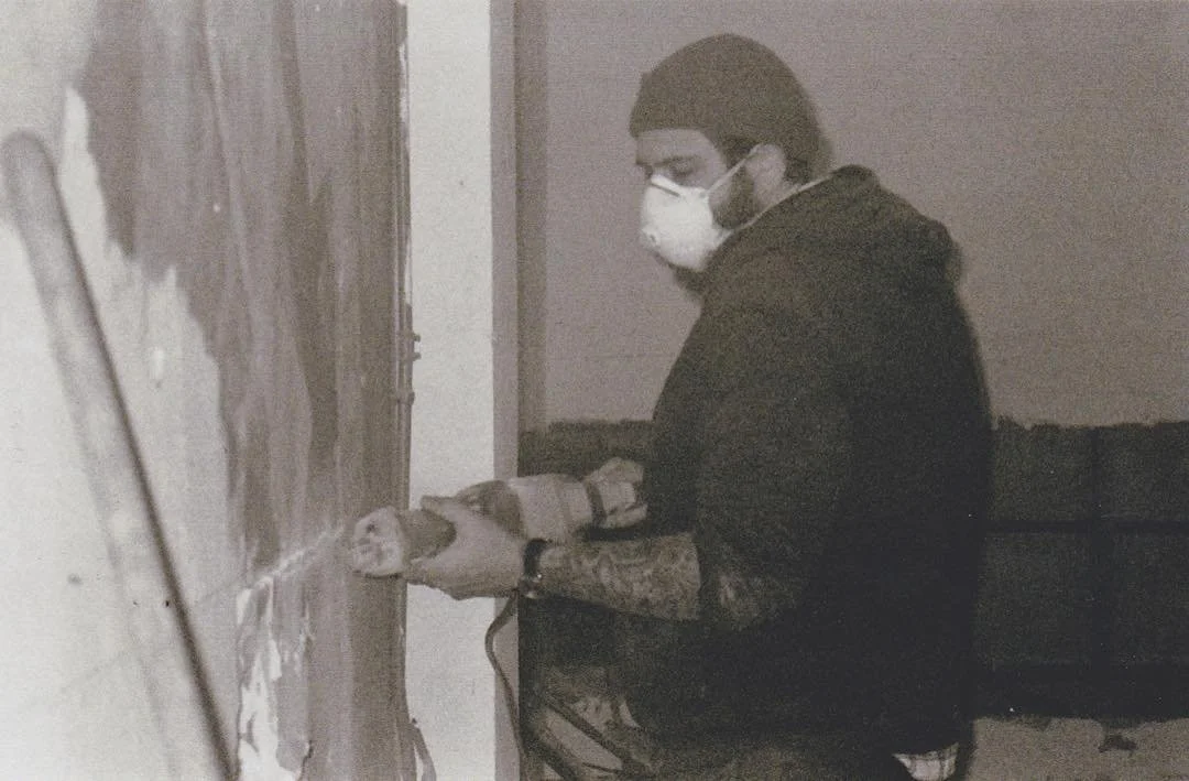 A person wearing a face mask, glasses, a beanie, and a tattooed arm is using a power tool to sand or paint a wall.
