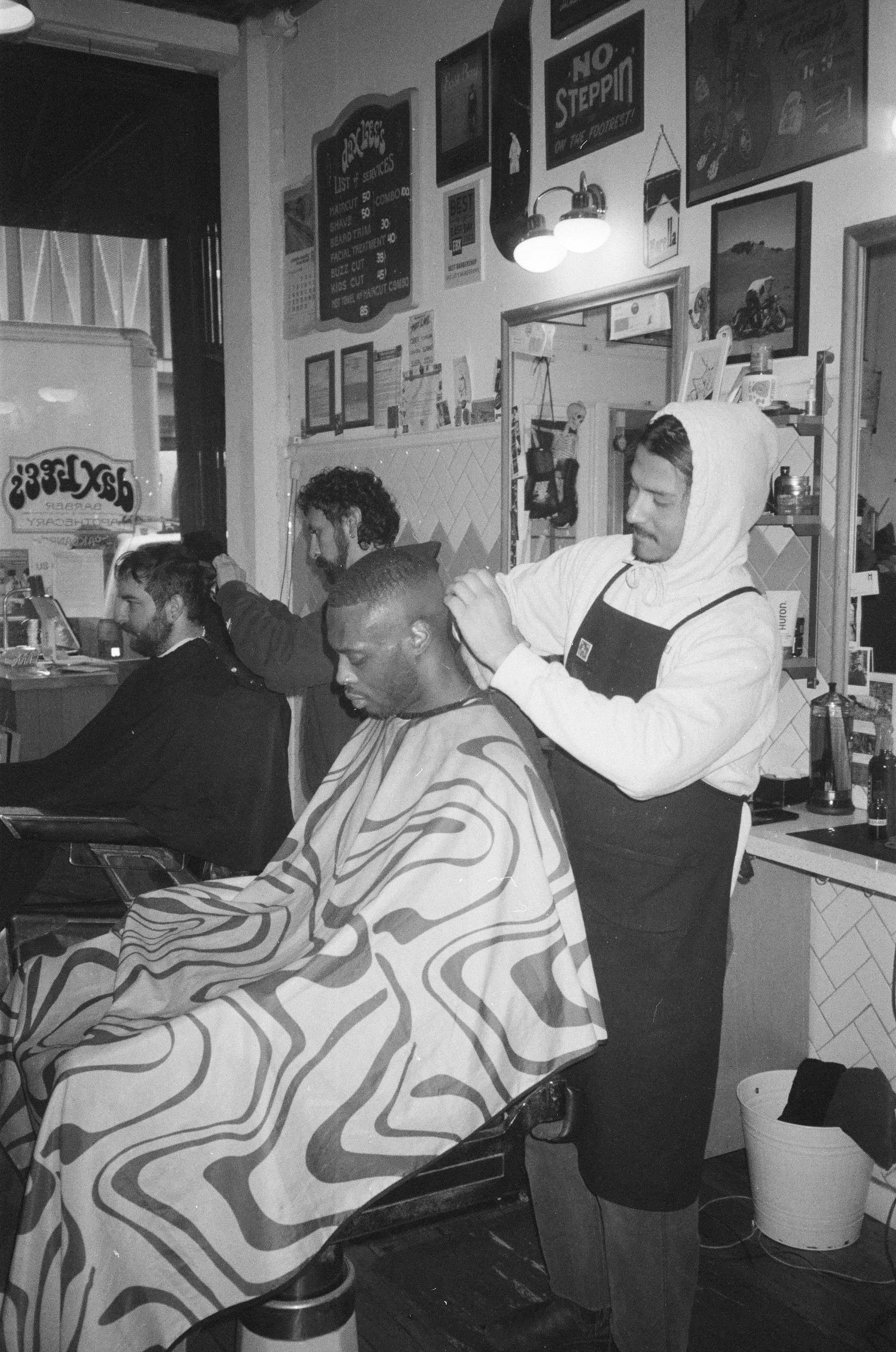 The photo shows a barber giving a haircut to a male customer in a barbershop, with two other customers seated and waiting. The walls are decorated with various posters and pictures, and there's a mirror behind the barber. The barber is wearing a hood