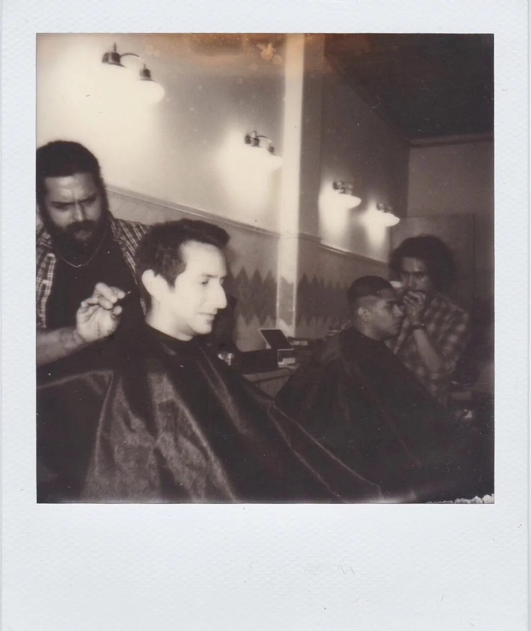 A man getting a haircut at a barbershop while two other men sit in chairs, one being the barber and the other across from him.