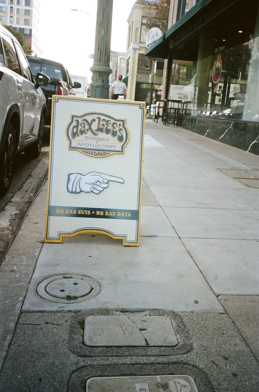 A sidewalk sign advertising Dax's Barber & Apothecary in Oakland, featuring a pointing hand illustration and the slogan 'No Bad Cuts - No Bad Days,' with parked cars on the street and buildings in the background.