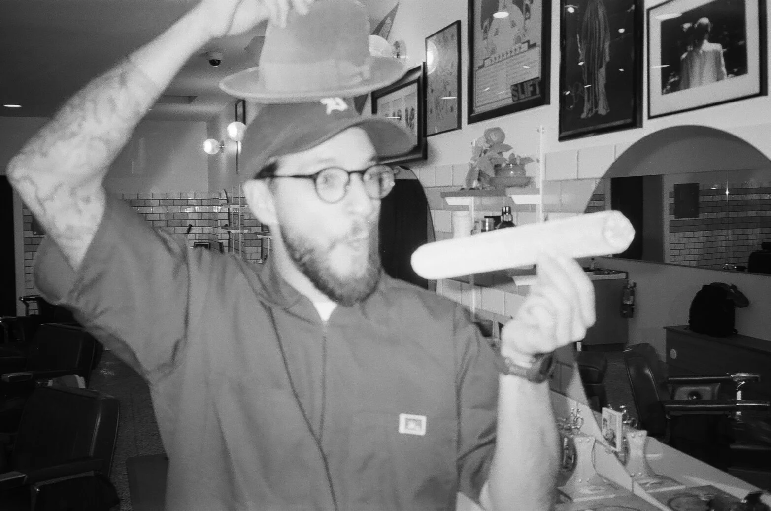 A man with glasses and a beard, wearing a cap, holds a Viking-style helmet with both hands and appears to be taking it off or putting it on in a casual indoor setting decorated with framed artworks.