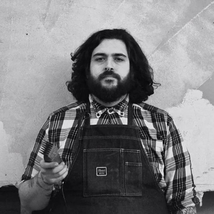 Black and white photo of a man with long curly hair and a beard, wearing a checkered shirt and apron, holding a comb in his right hand, standing in front of a textured wall.