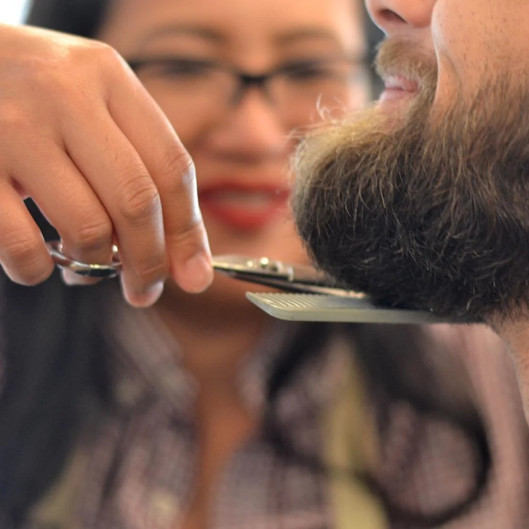 A hairstylist trims a man's beard using scissors and a comb, with a woman smiling and watching in the background.