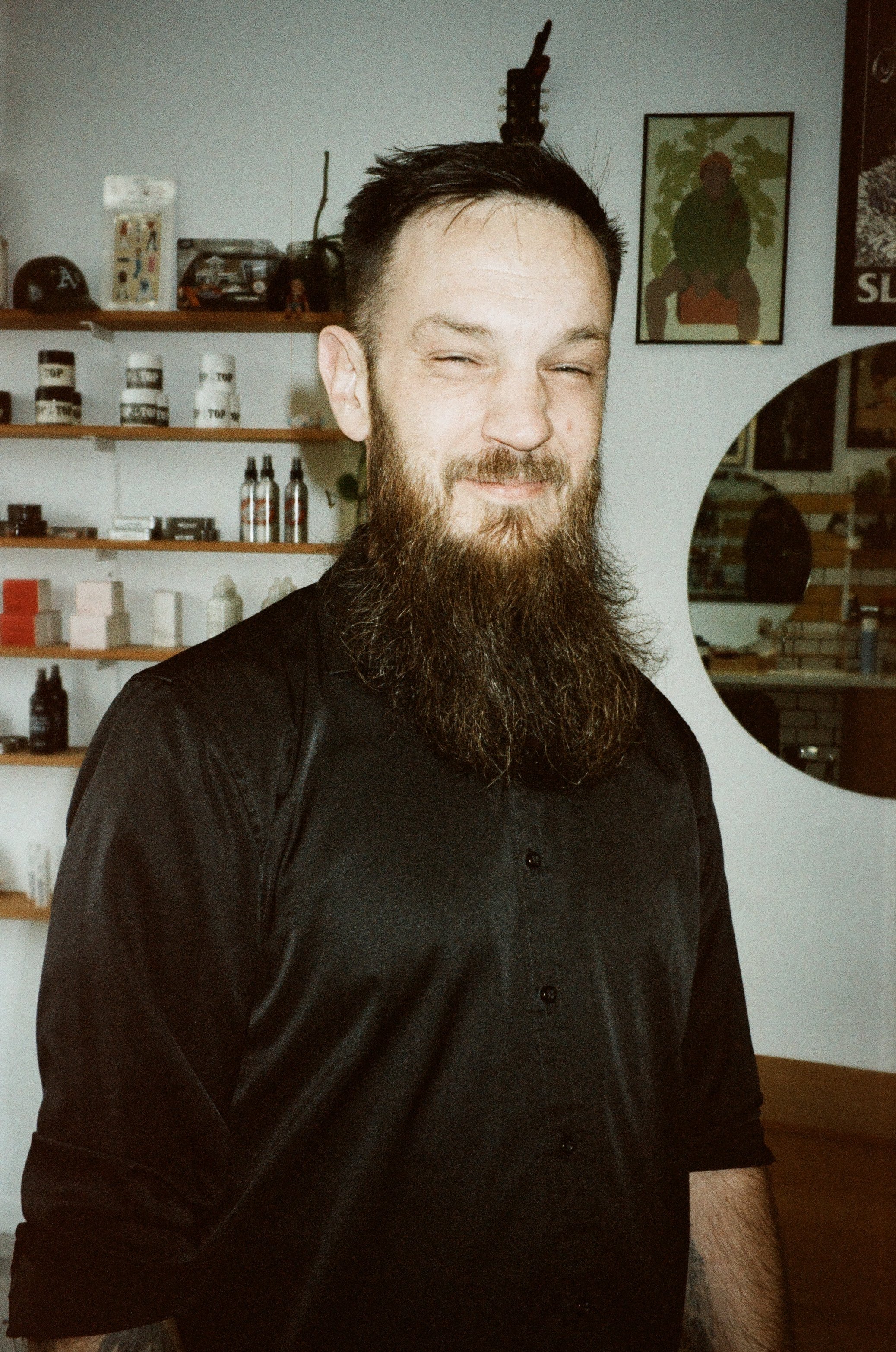 A man with a beard and short dark hair wearing a black shirt, smiling with eyes partially closed, standing in a room with shelves filled with various items including bottles and model cars, and framed artwork on the wall.