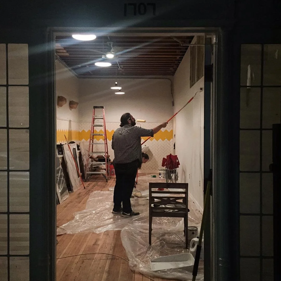 A man painting a wall with a roller, standing on the floor inside a room under renovation, with a ladder and painting supplies around.