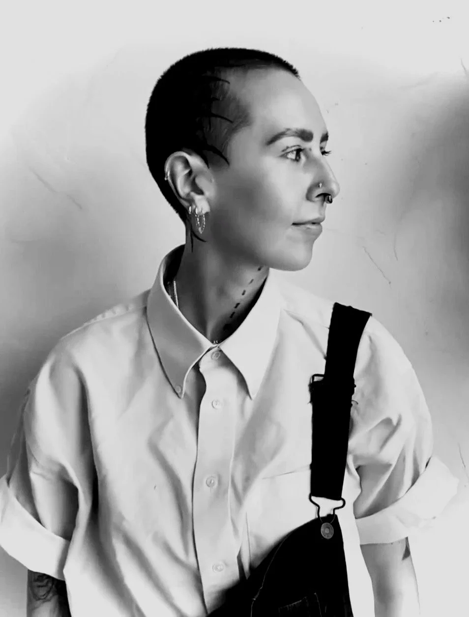 A black and white portrait of a young woman with short hair, multiple facial piercings, earrings, and a nose ring, wearing a white button-up shirt and black overalls.