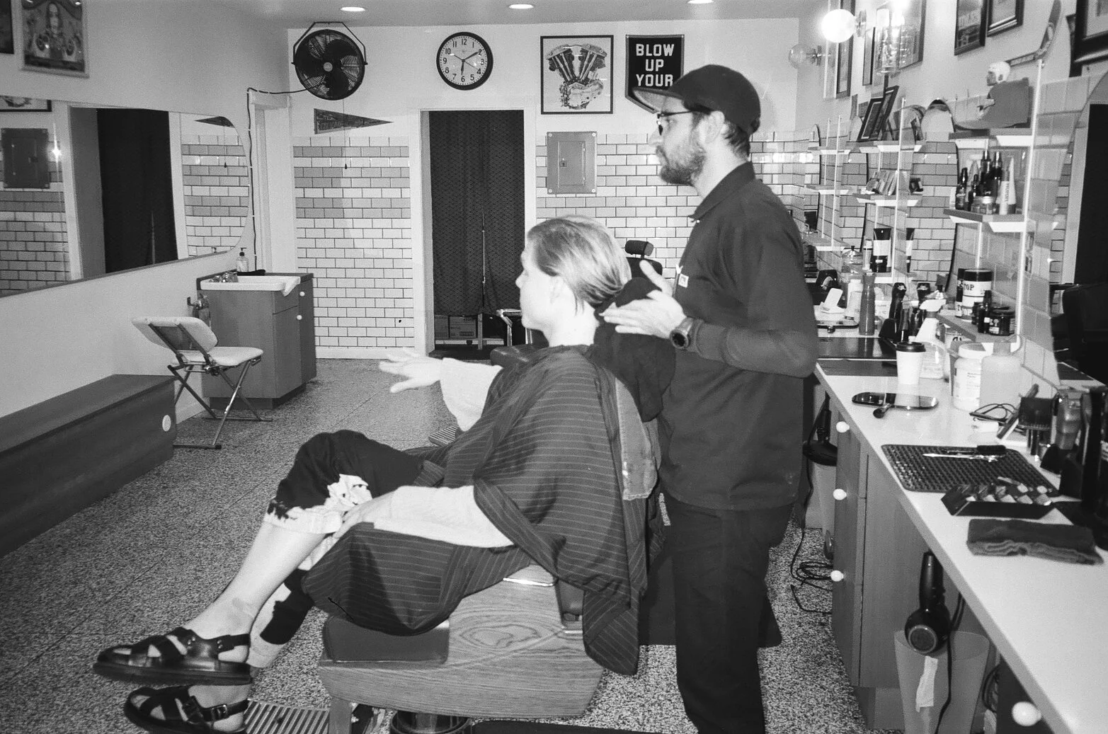 A person is getting a haircut at a barbershop while a barber stands behind them, holding scissors. The barbershop has a brick wall, a clock, a wall fan, and shelves with barber supplies.