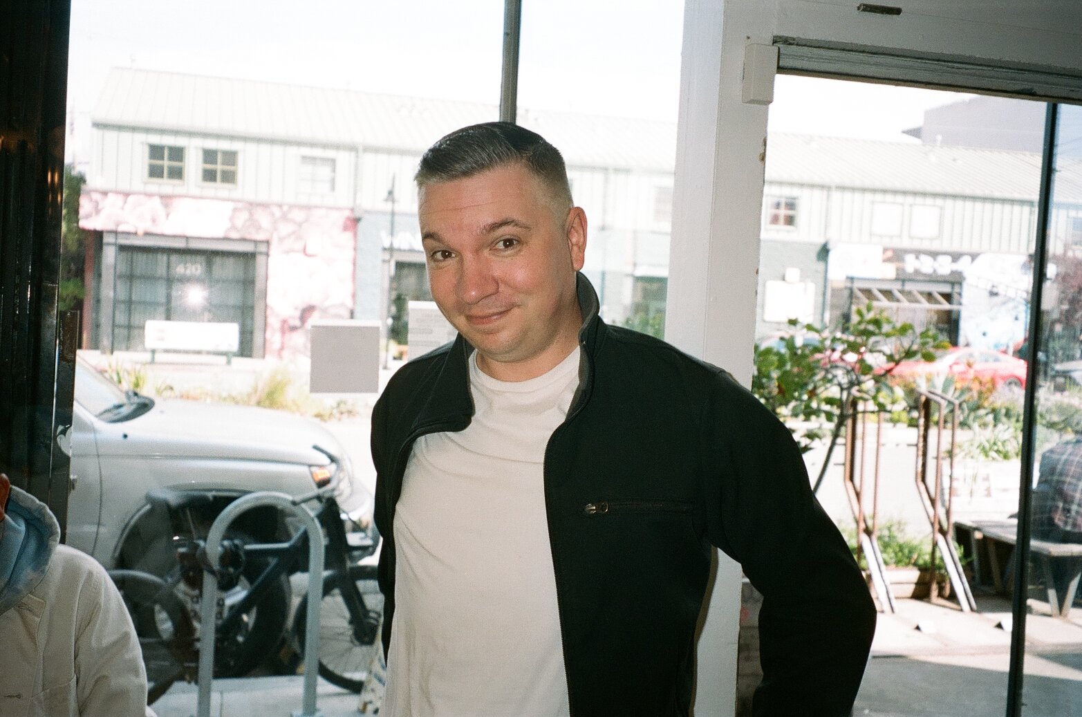 A man with short dark hair, wearing a white t-shirt and black jacket, standing indoors near a window, smiling at the camera.