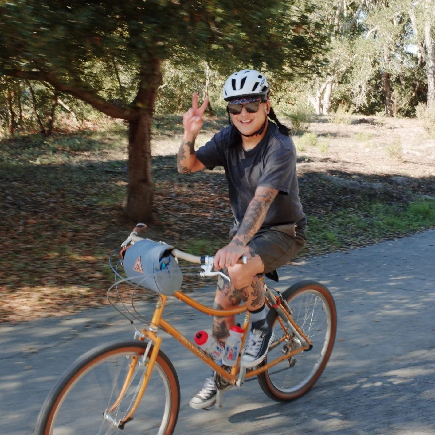 A happy person with tattoos, wearing a white helmet, sunglasses, and a black shirt, riding an orange bicycle on a trail surrounded by trees, making a peace sign with their right hand.