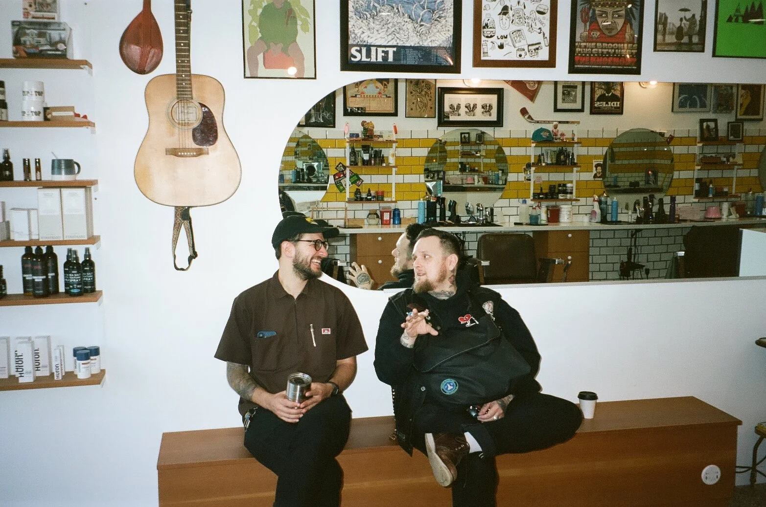 Two men sitting on a wooden bench in a cafe, smiling and talking to each other. One is wearing a brown shirt and glasses, holding a can, and the other is dressed in black with tattoos and making a gesture with his hand. Behind them is a mirror with a