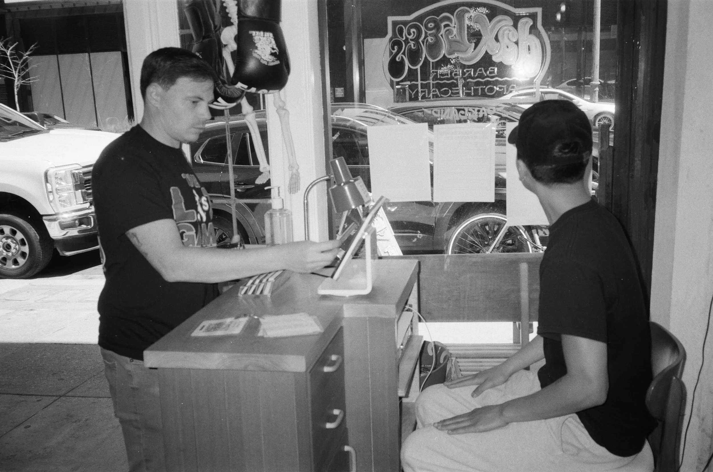 A young man is sitting at a table inside a pizza restaurant, wearing a black shirt and a baseball cap. An older man is standing nearby, reading from a menu or pamphlet. The window behind them has a neon sign that reads "Pizza" and "Pizzeria". Outside