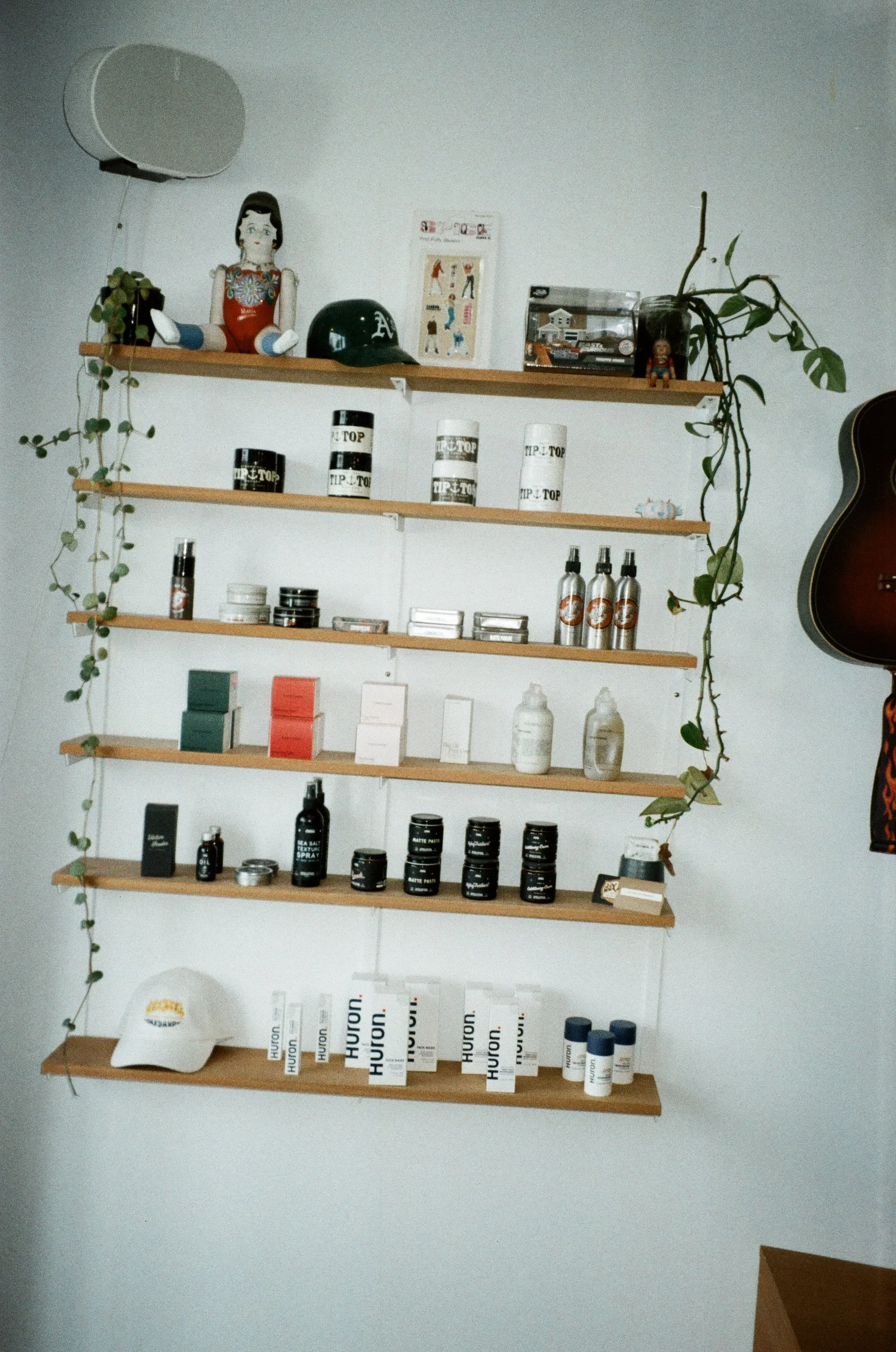 Wall-mounted wooden shelves displaying various branded products including cans and bottles, a toy doll, a black football helmet, decorative items, and a guitar on the right side. Green trailing plants decorate the shelves.
