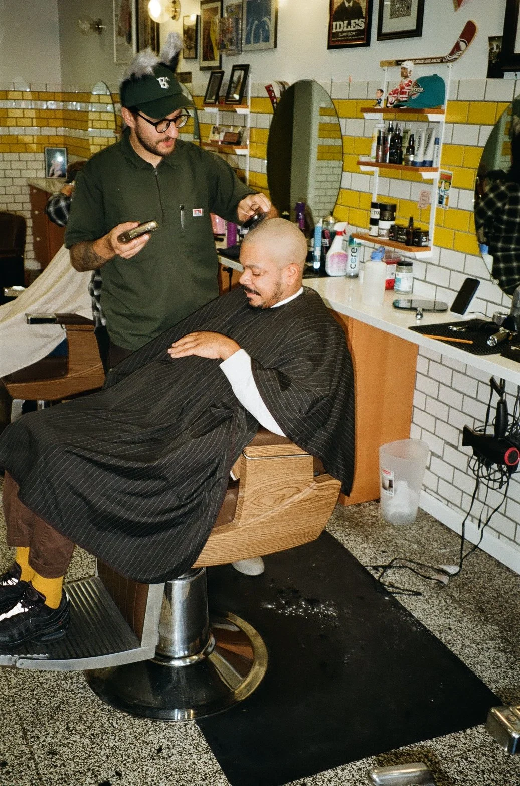 A man with a shaved head and beard sitting in a barber chair, having his hair cut or styled by a barber in a barbershop. The barber is wearing glasses and a green shirt and is holding a phone. The barbershop has tiled walls, mirrors, shelves with hai