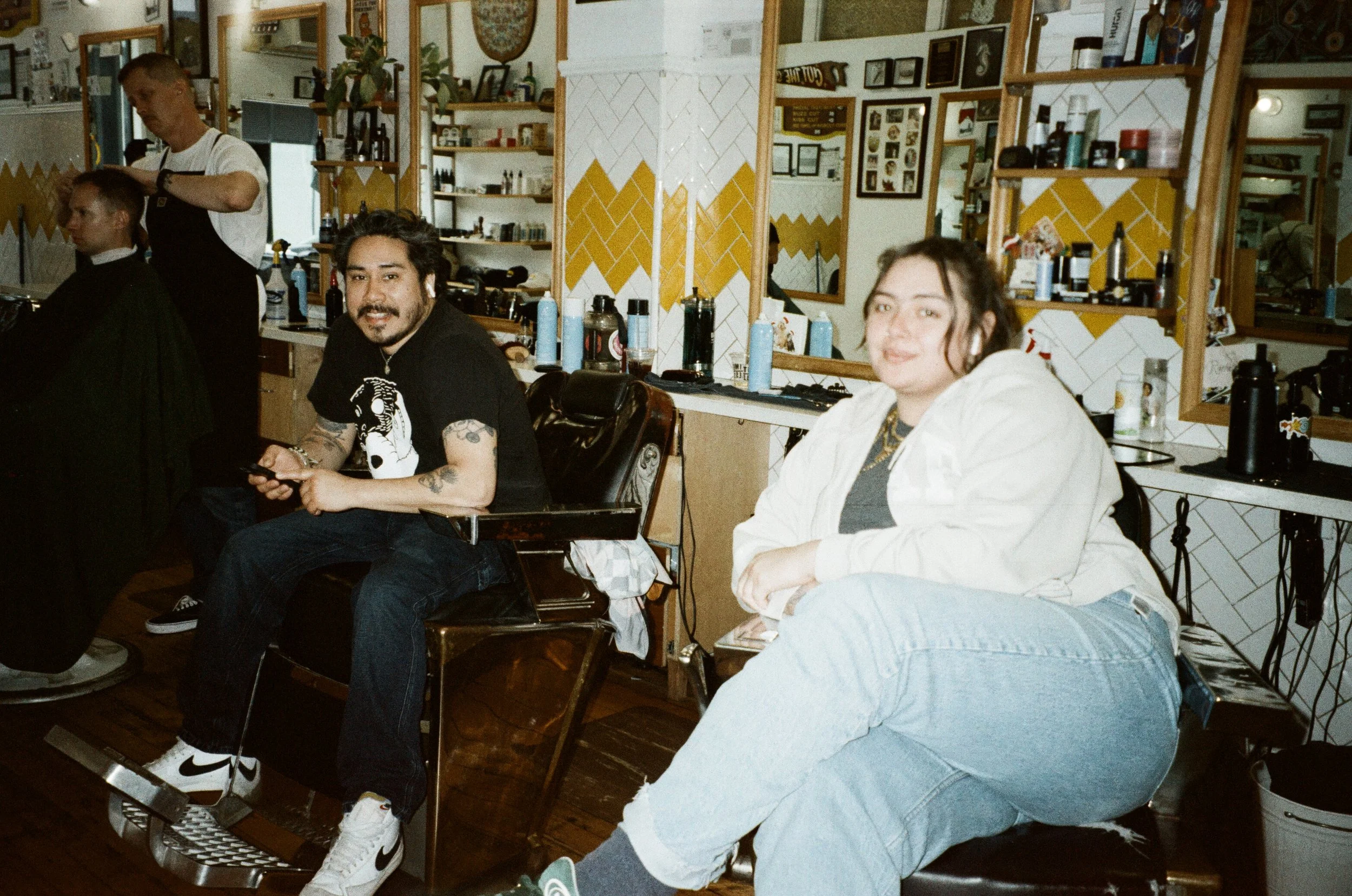 People in a barbershop receiving haircuts, with a barber cutting a man's hair on the left, and a woman sitting nearby in the foreground. The shop has yellow and white tiled walls, mirrors, and shelves with hair products.
